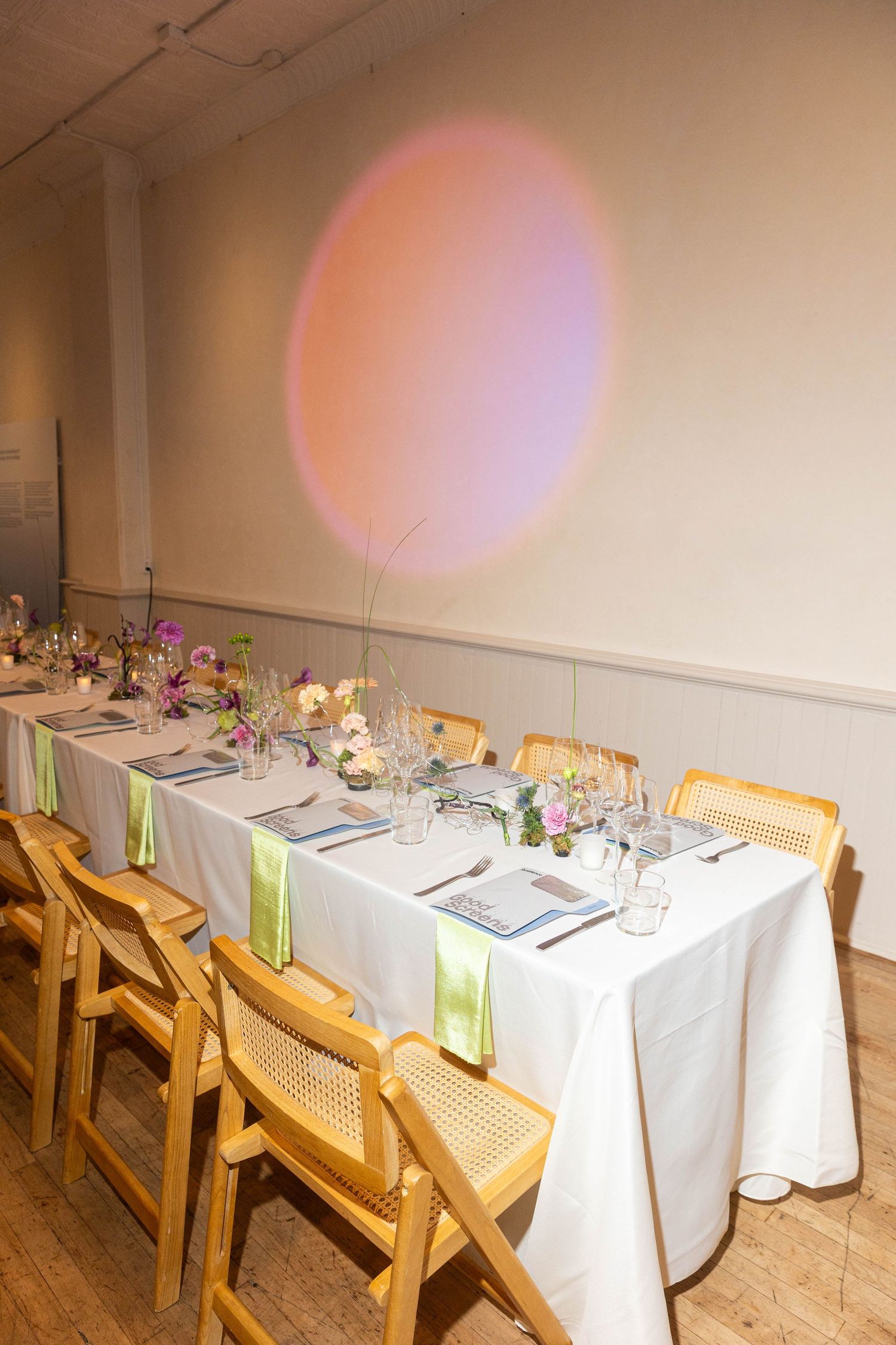 Long banquet table with white linens, green napkins, wooden chairs, and fresh floral centerpieces beneath a soft pink spotlight on the wall.