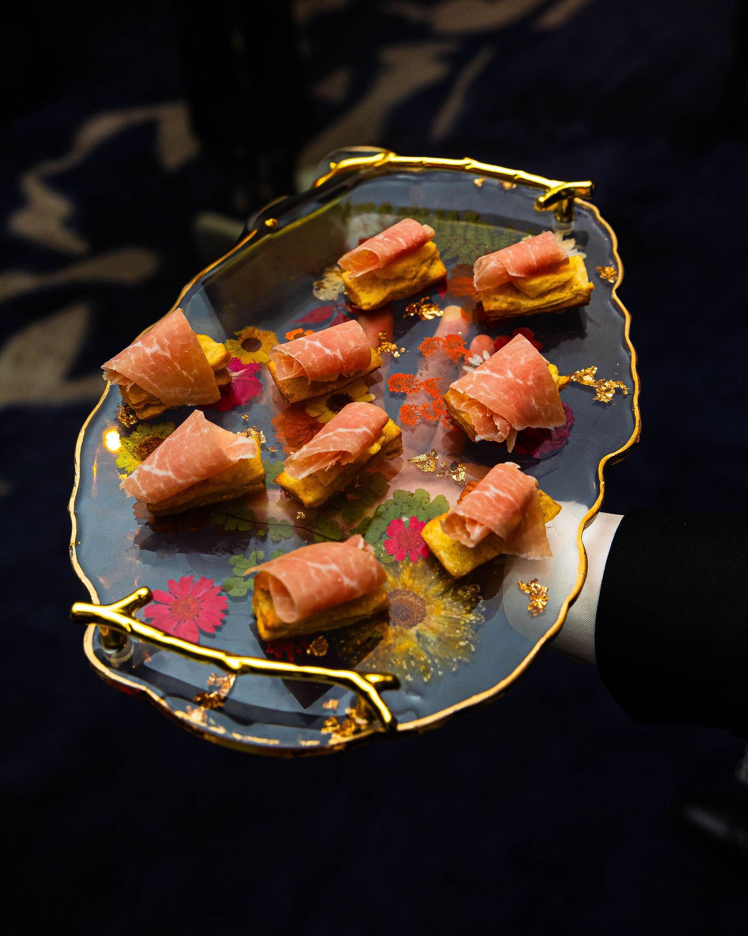 Waiter holding an ornate floral serving tray with prosciutto-wrapped canapés on golden toasted bites.