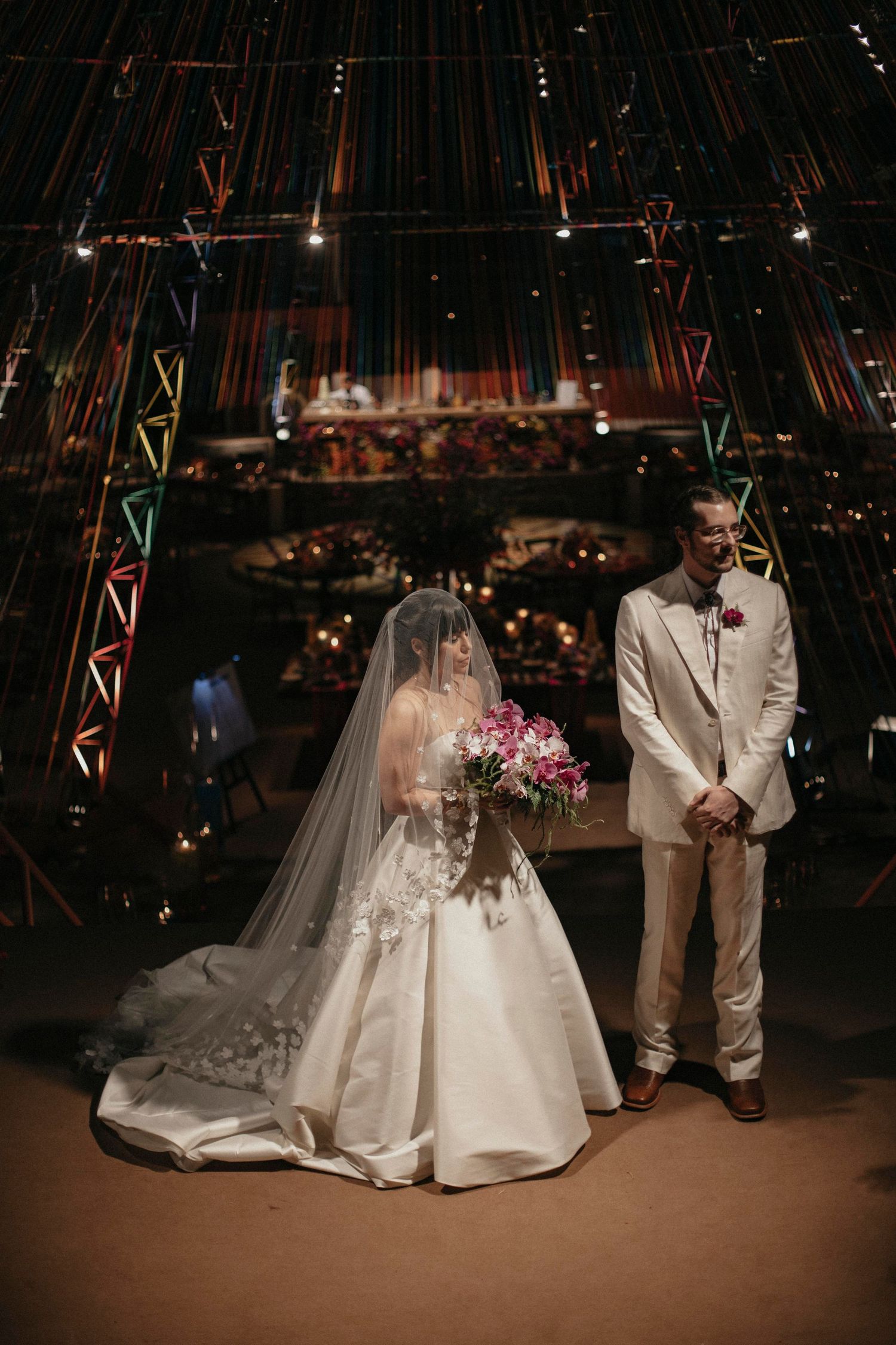 Bride in ballgown with veil holding pink bouquet stands beside groom at colorful wedding venue.