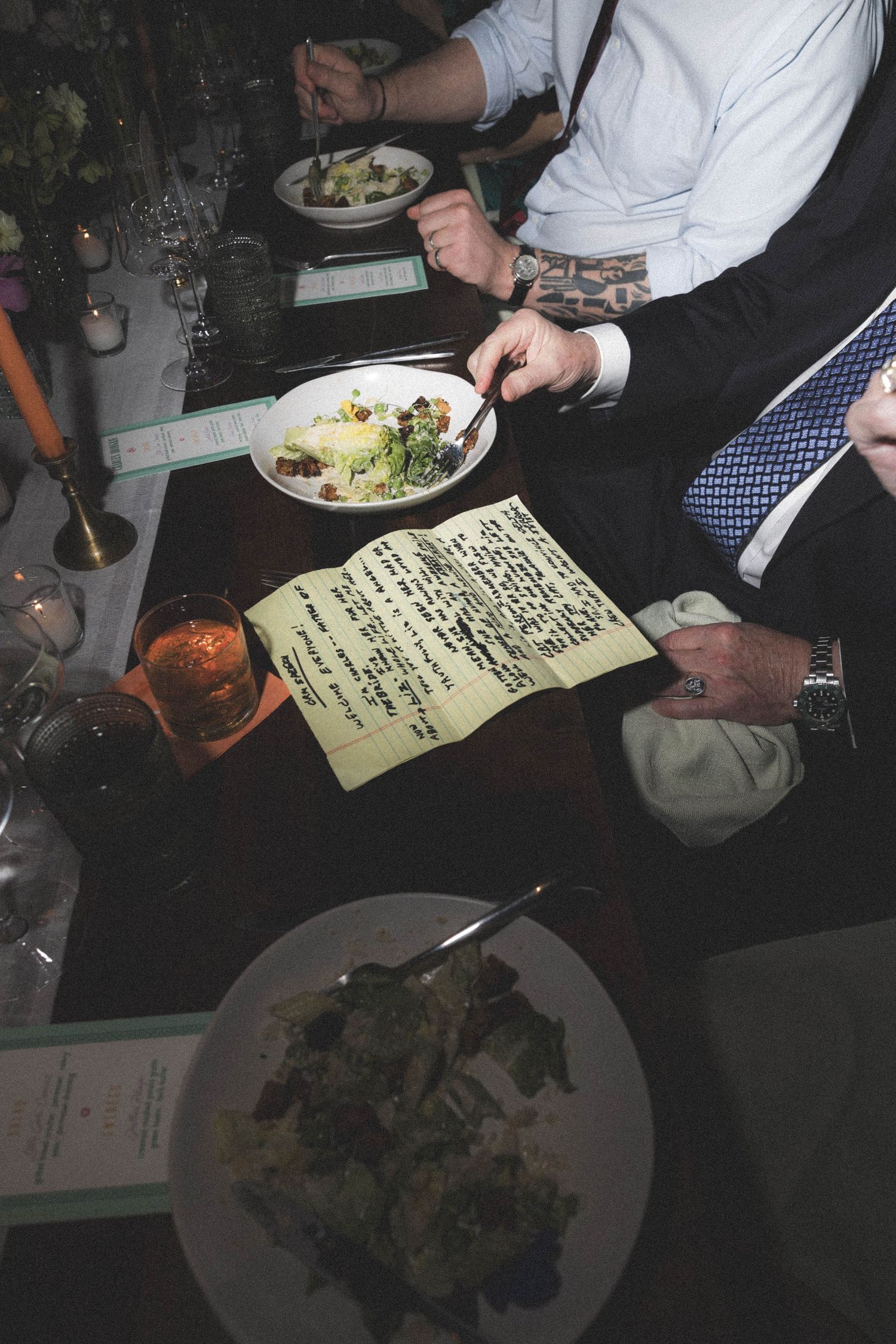 Wedding guests review handwritten notes beside salad plates at a candlelit reception dinner.