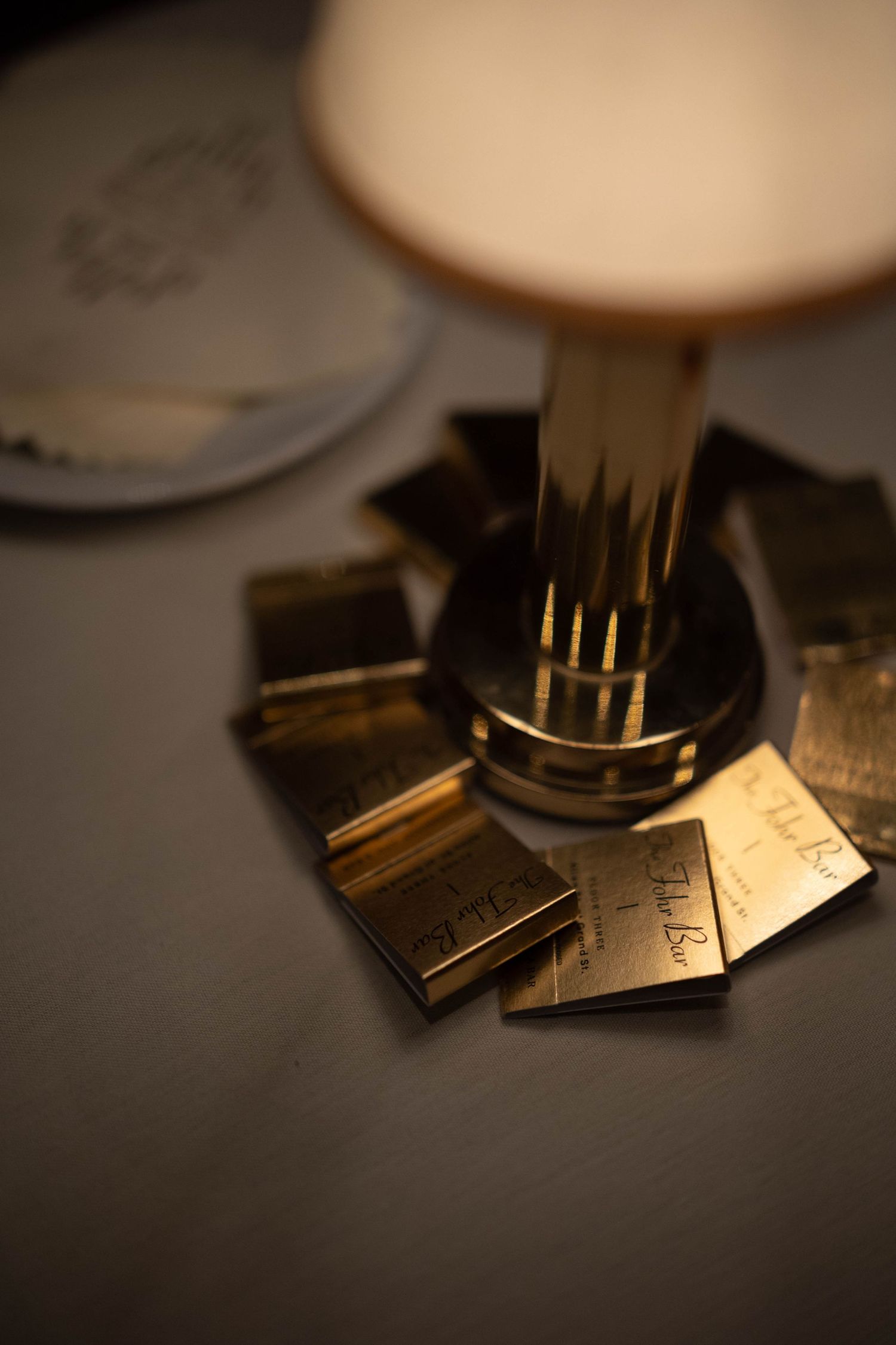 Gold mirrored acrylic place cards with elegant script lettering arranged around the base of a glowing table lamp on a dimly lit table.