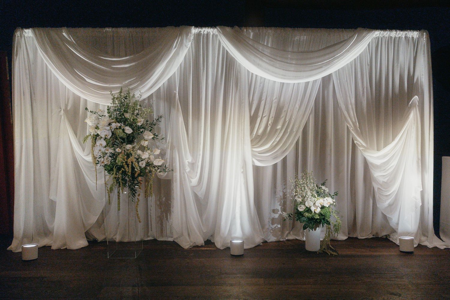 Elegant white draped fabric backdrop with floral arrangements and uplighting on a stage, ideal for wedding ceremonies and event decor.