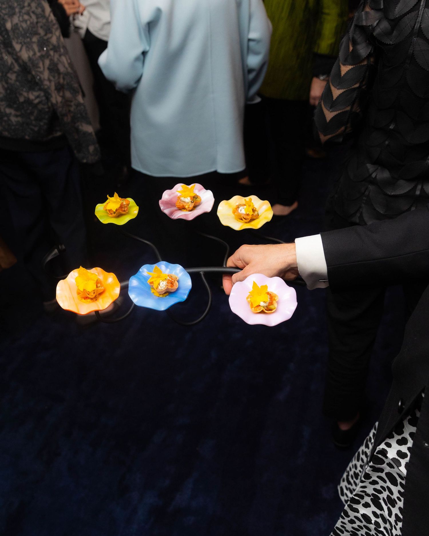 A hand holds a tray of colorful flower-shaped dishes with glowing orange centers while guests in formal attire stand on a dark blue carpet.