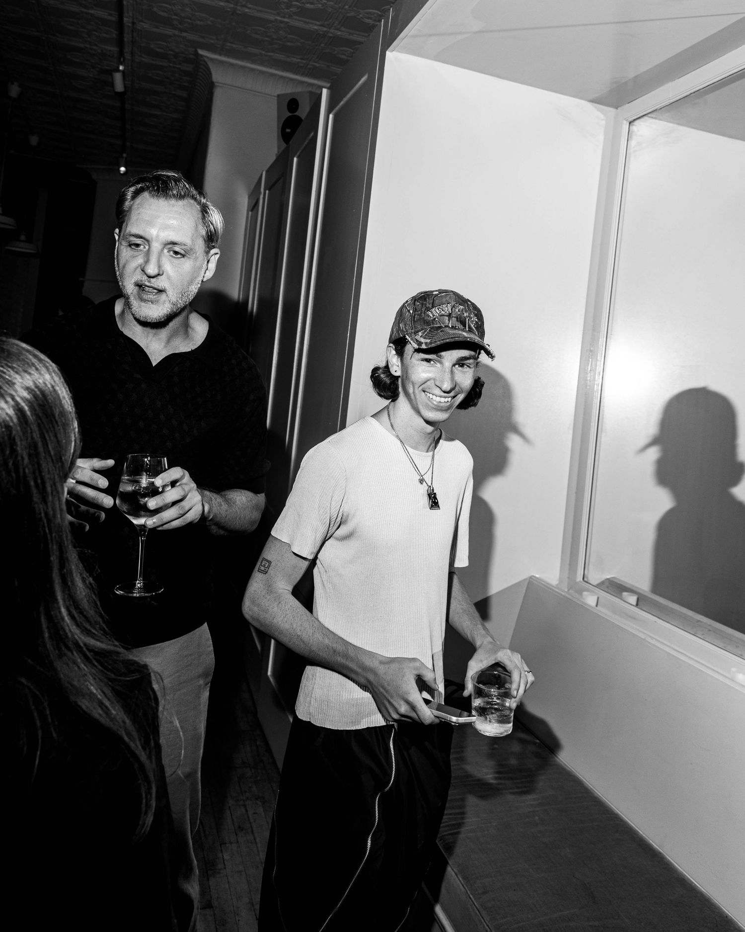 Black and white photo of a smiling young man in a patterned cap carrying drinks past a man holding a wine glass at a lively party.