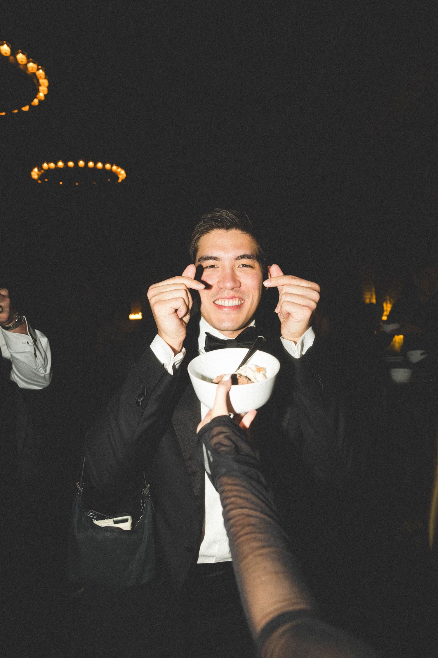 Smiling young man in tuxedo adjusting bow tie at dark wedding reception venue.