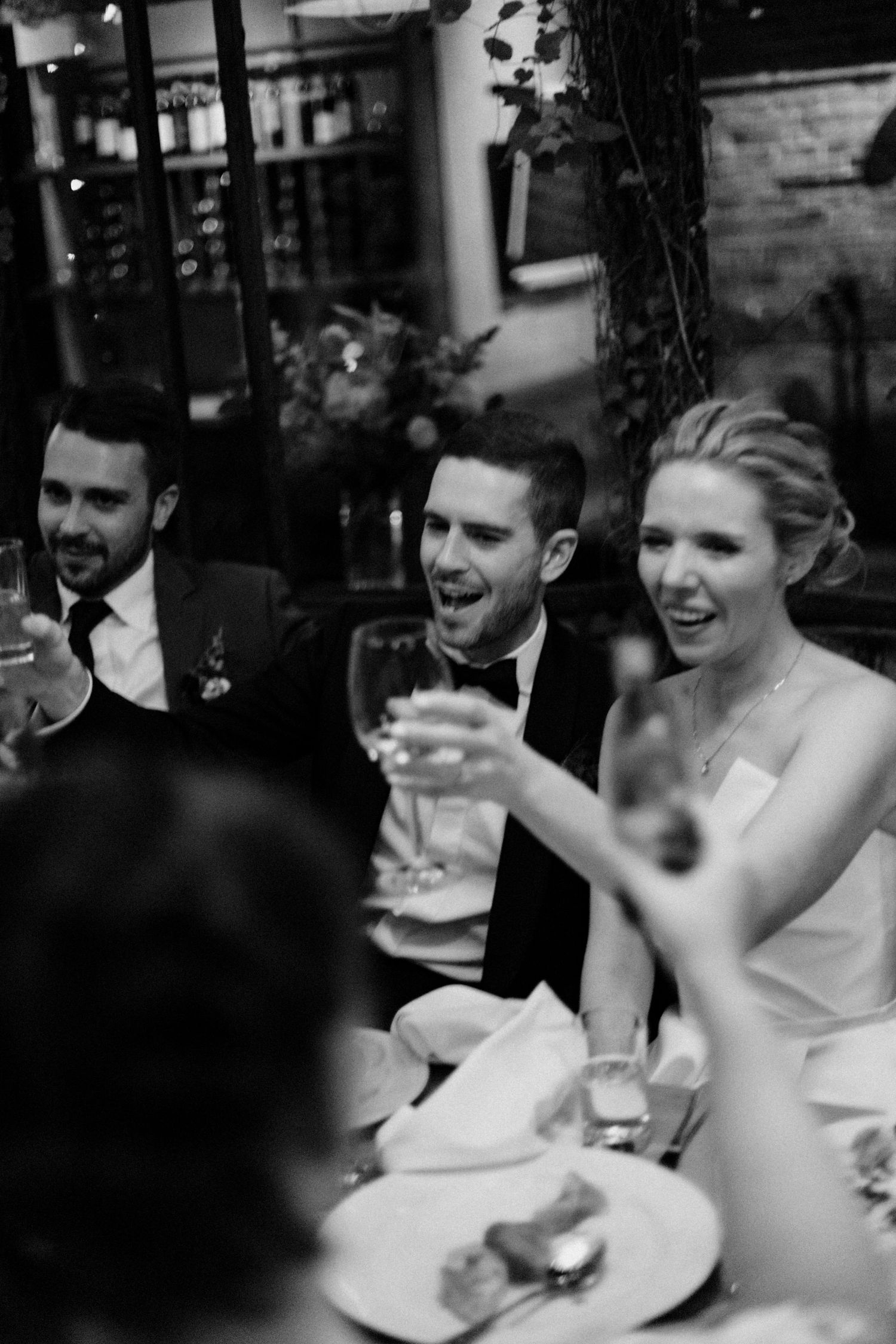 Black and white photo of bride and groom laughing and toasting with champagne glasses at wedding reception dinner.
