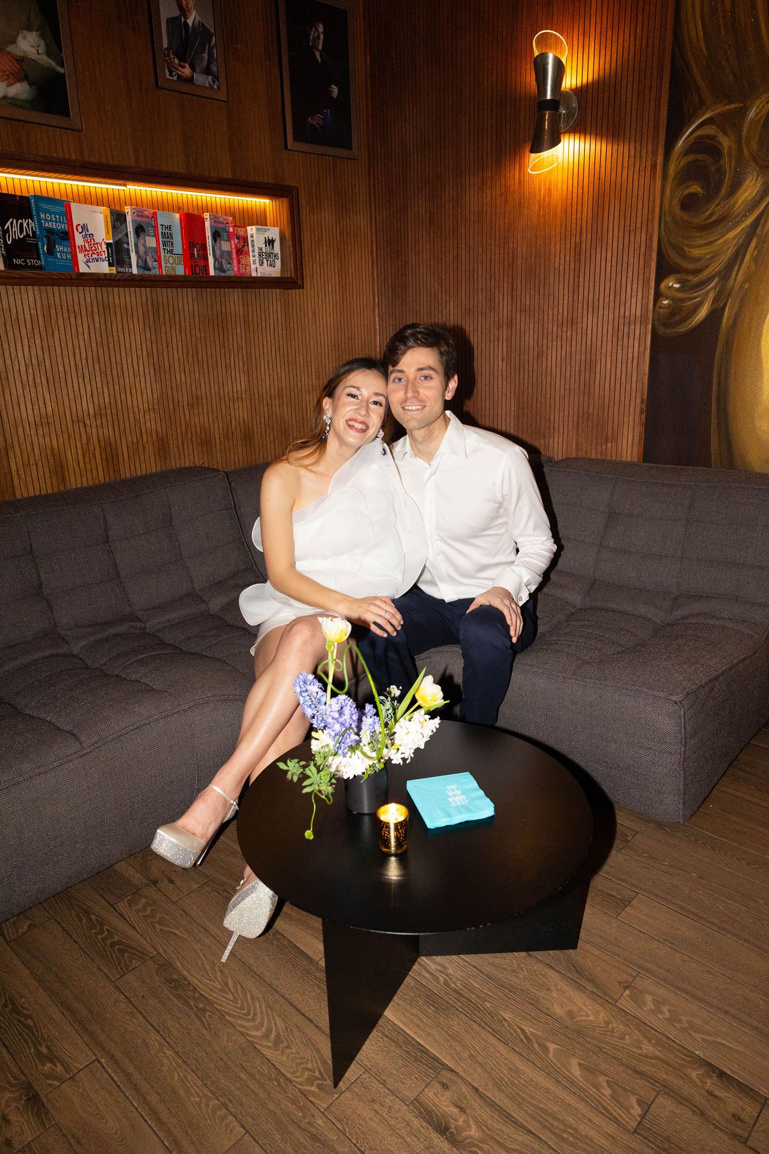 Smiling couple in white outfits sit together on gray sofa with flowers on coffee table at restaurant.