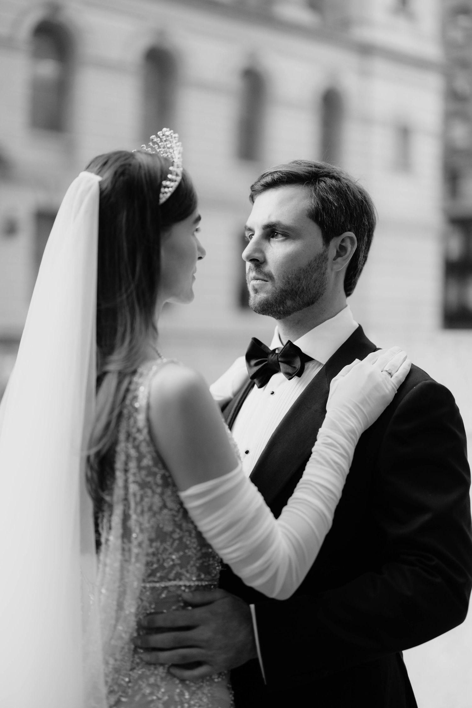 Black and white portrait of bride in beaded gown and tiara embracing groom in tuxedo on a city street.