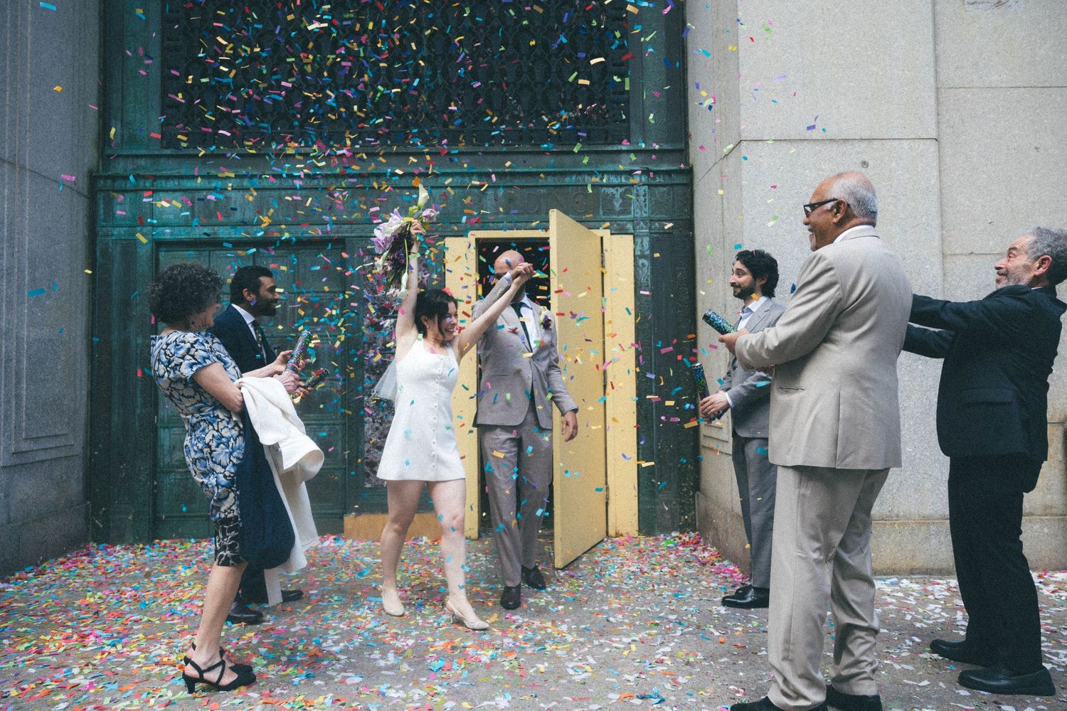 Bride and groom celebrating with colorful confetti shower outside with small group of guests.