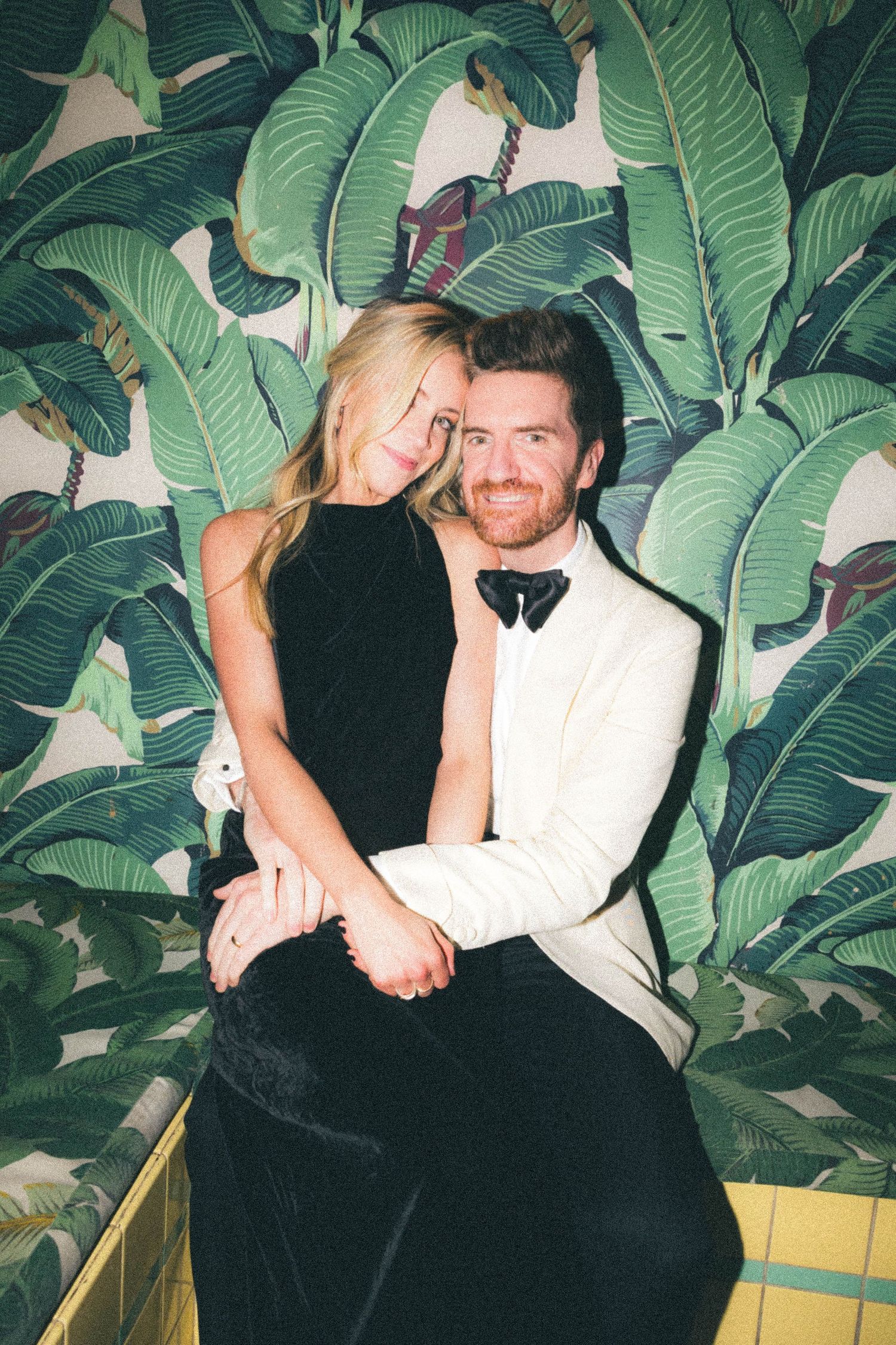 Smiling couple posing together in front of tropical banana leaf wallpaper at event.