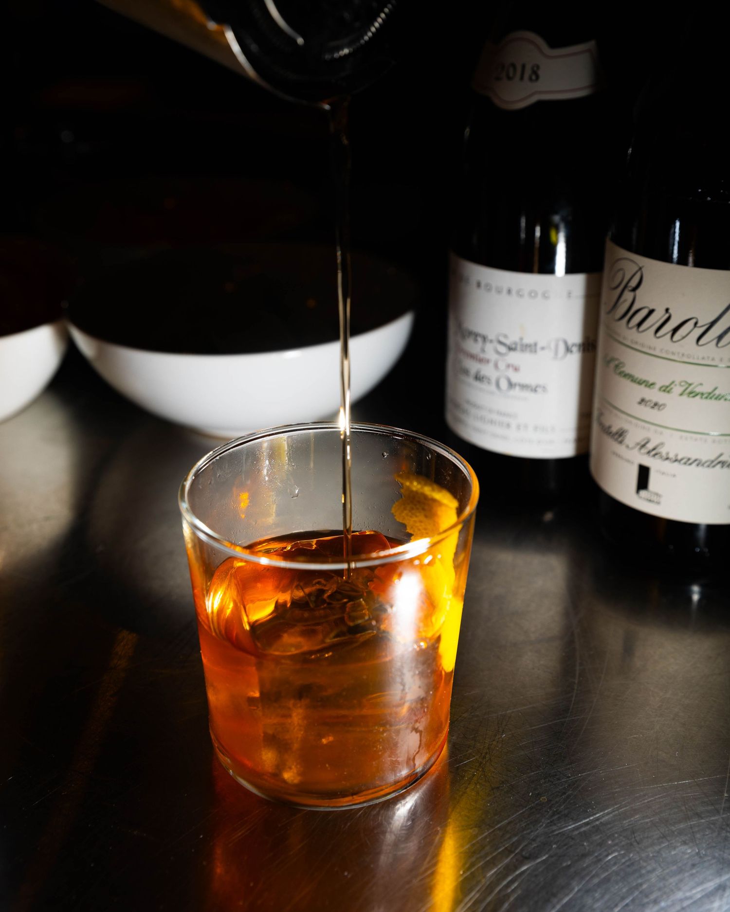 Amber liquid being poured into a glass with ice on a stainless steel bar, with Burgundy and Barolo wine bottles in the background.