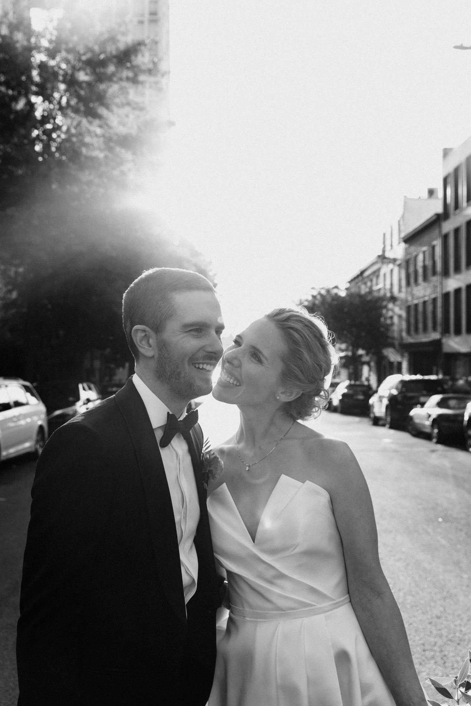 Smiling bride and groom in black tuxedo share joyful moment on sunny city street, black and white photo.