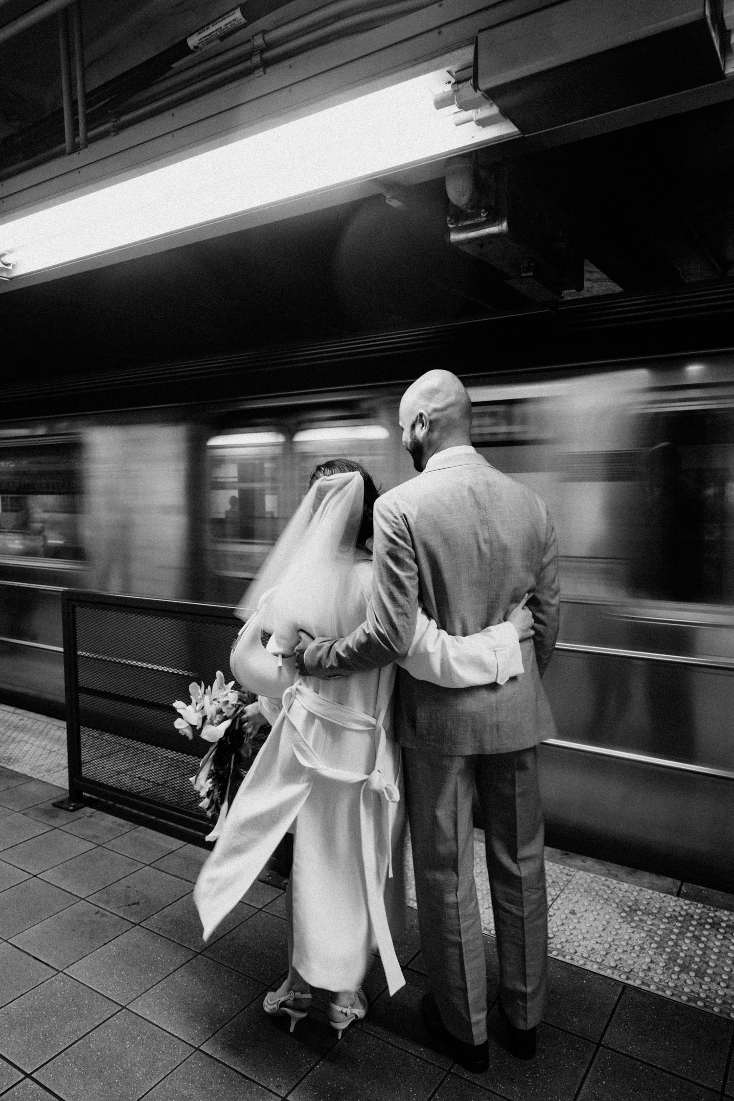 Black and white photo of newlyweds embracing on a subway platform as a train passes by.