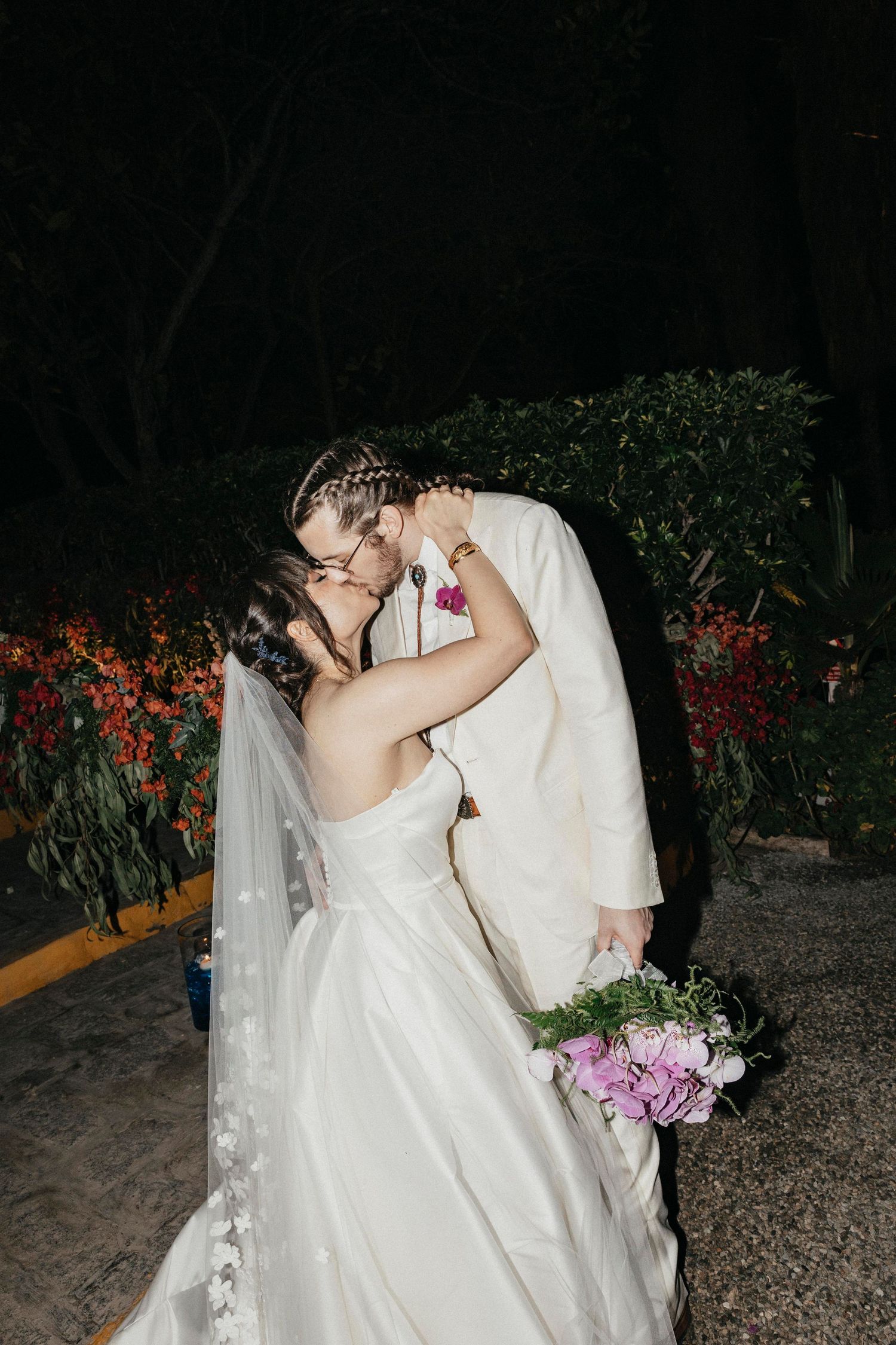 Bride holding purple bouquet shares a tender kiss with groom in cream suit amid garden wedding at night.