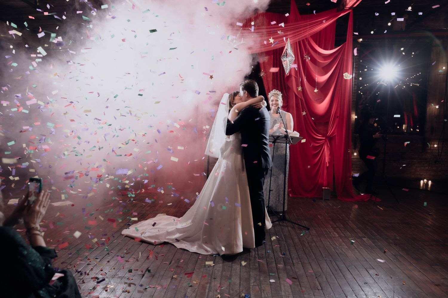 Bride and groom dance under colorful confetti with red curtain backdrop at elegant wedding reception.