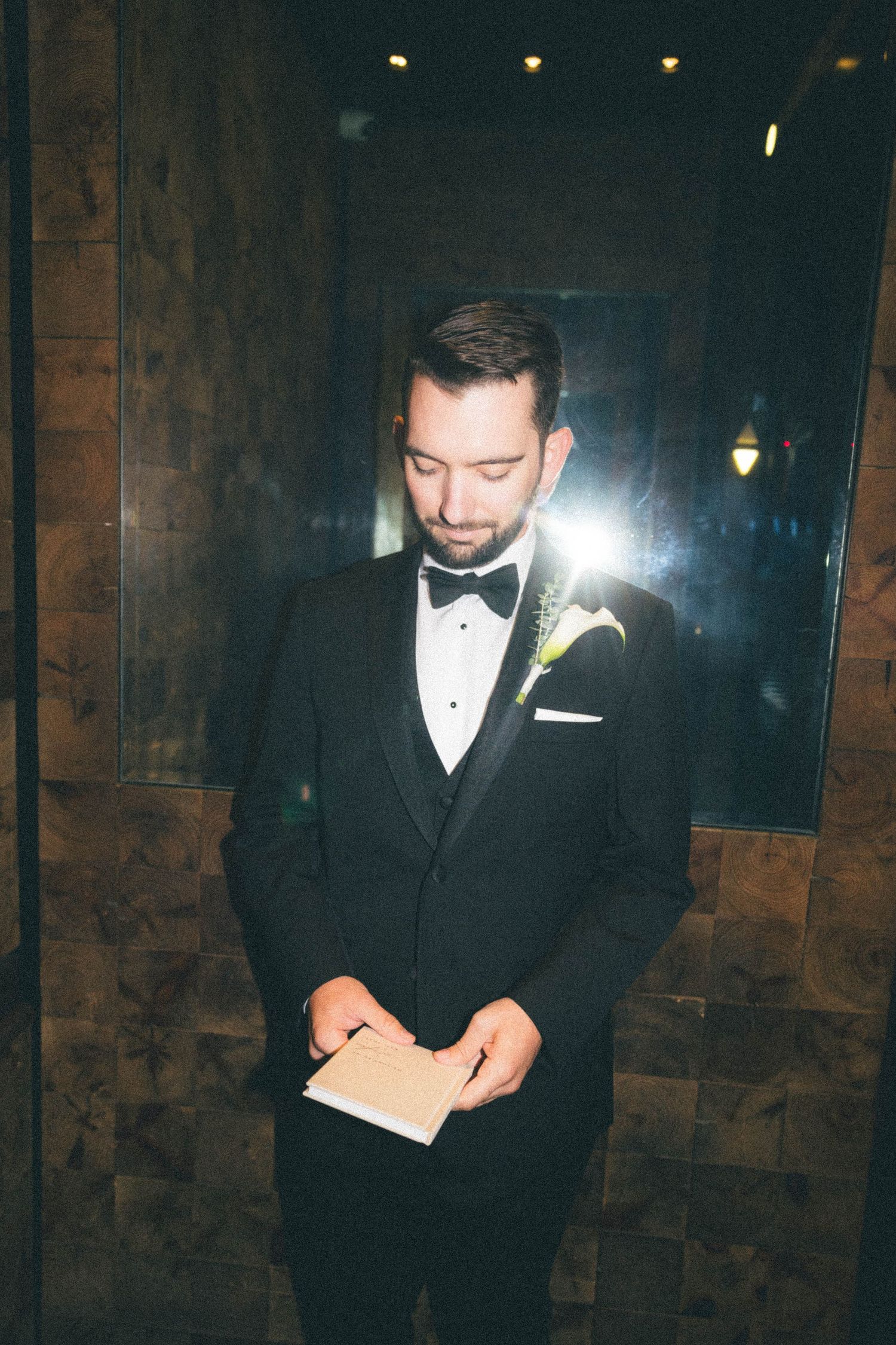 Groom in black tuxedo with boutonniere reading vows from small notebook in dimly lit venue.