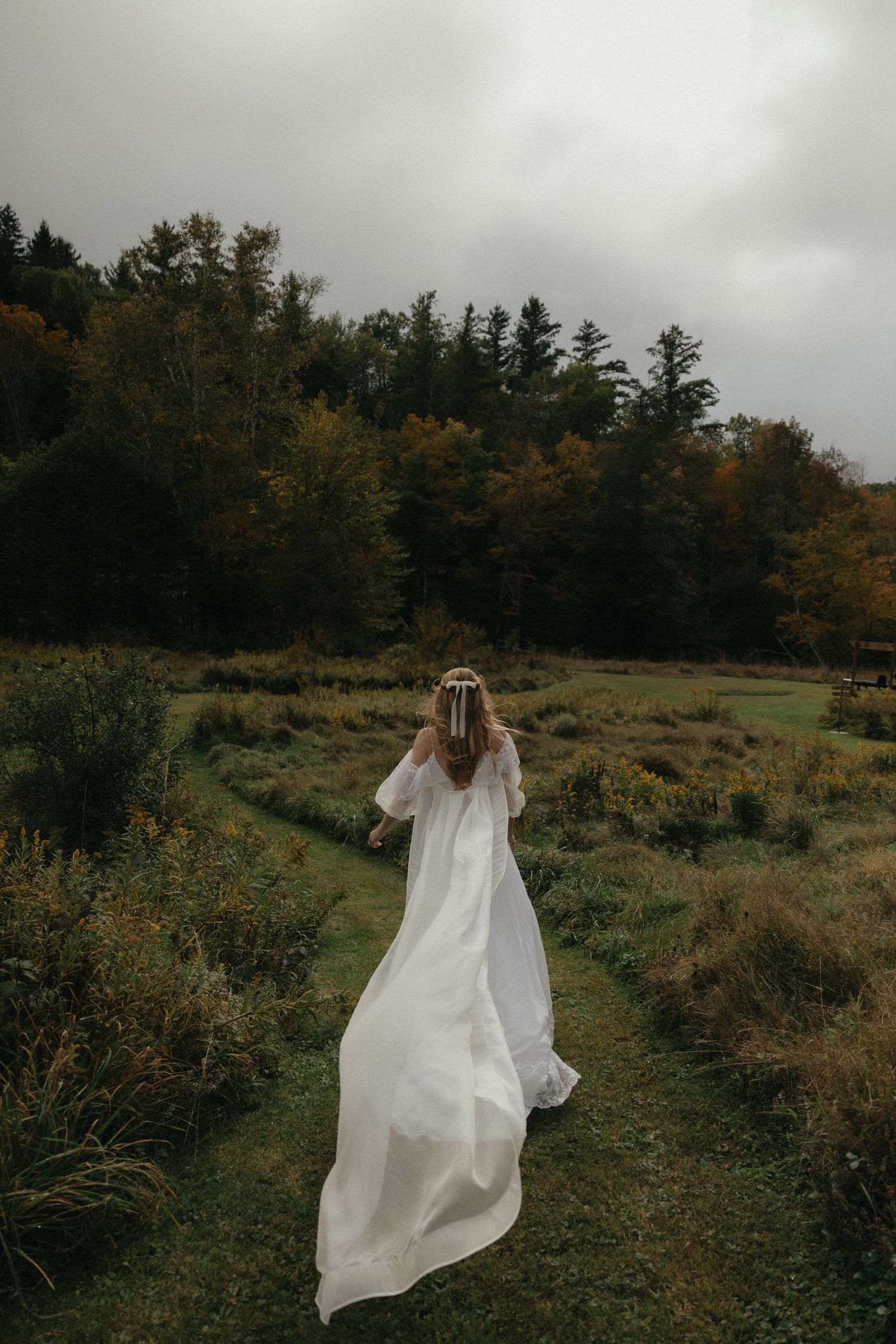 Bride in flowing white gown walking through a lush green meadow surrounded by autumn trees on her wedding day.