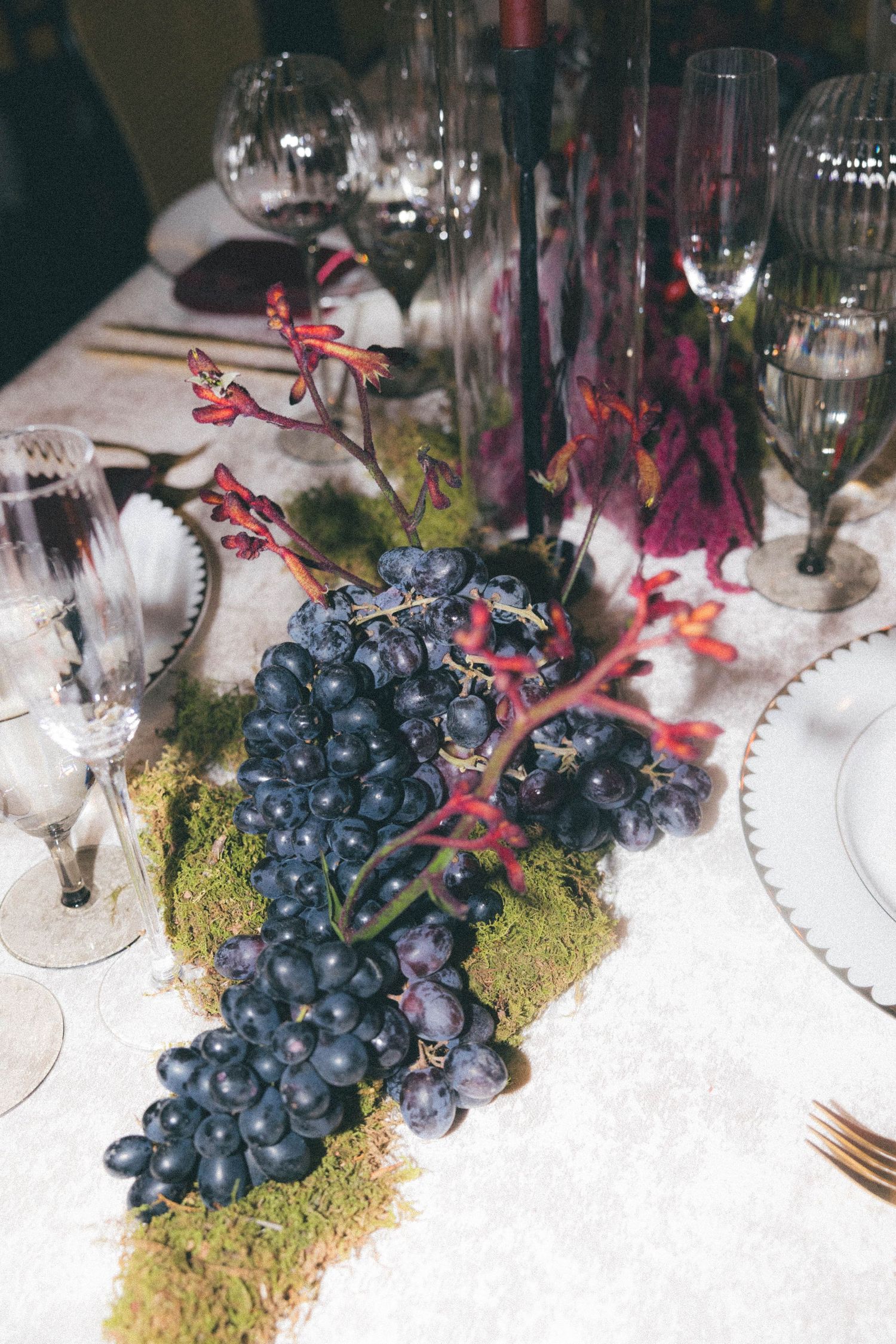 Elegant wedding table centerpiece featuring dark grapes, moss, and tall floral arrangements with crystal glassware.