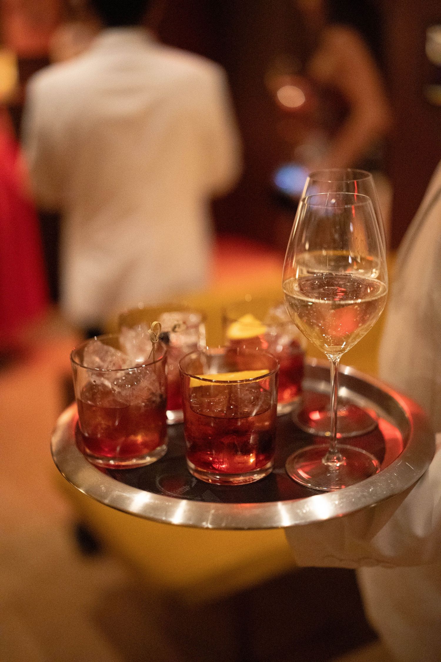 Server in white holding a silver tray with iced red cocktails garnished with citrus and a glass of white wine at an elegant event.