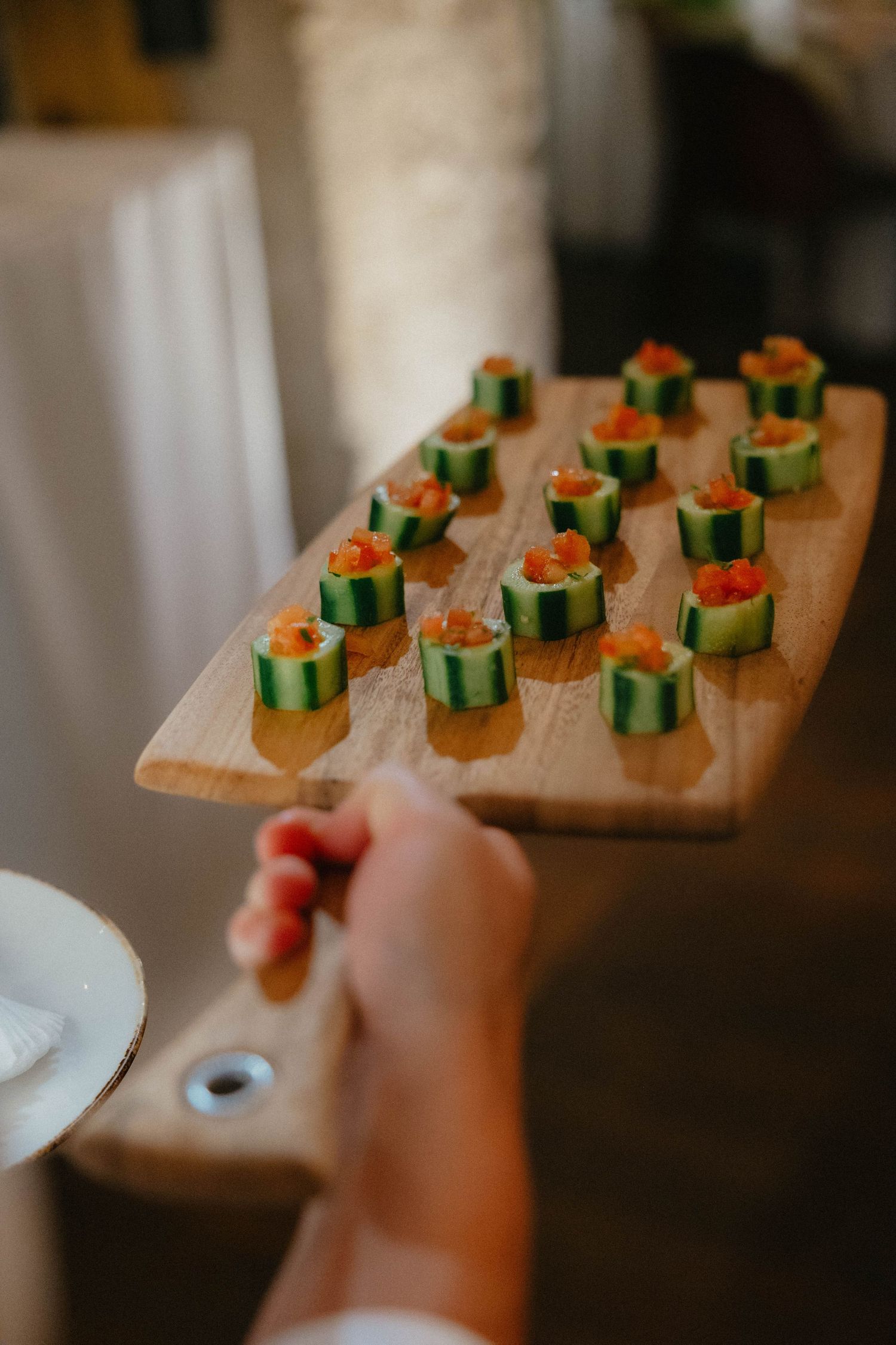 Hand holding a wooden serving board topped with cucumber cup appetizers filled with diced salmon at an elegant event.