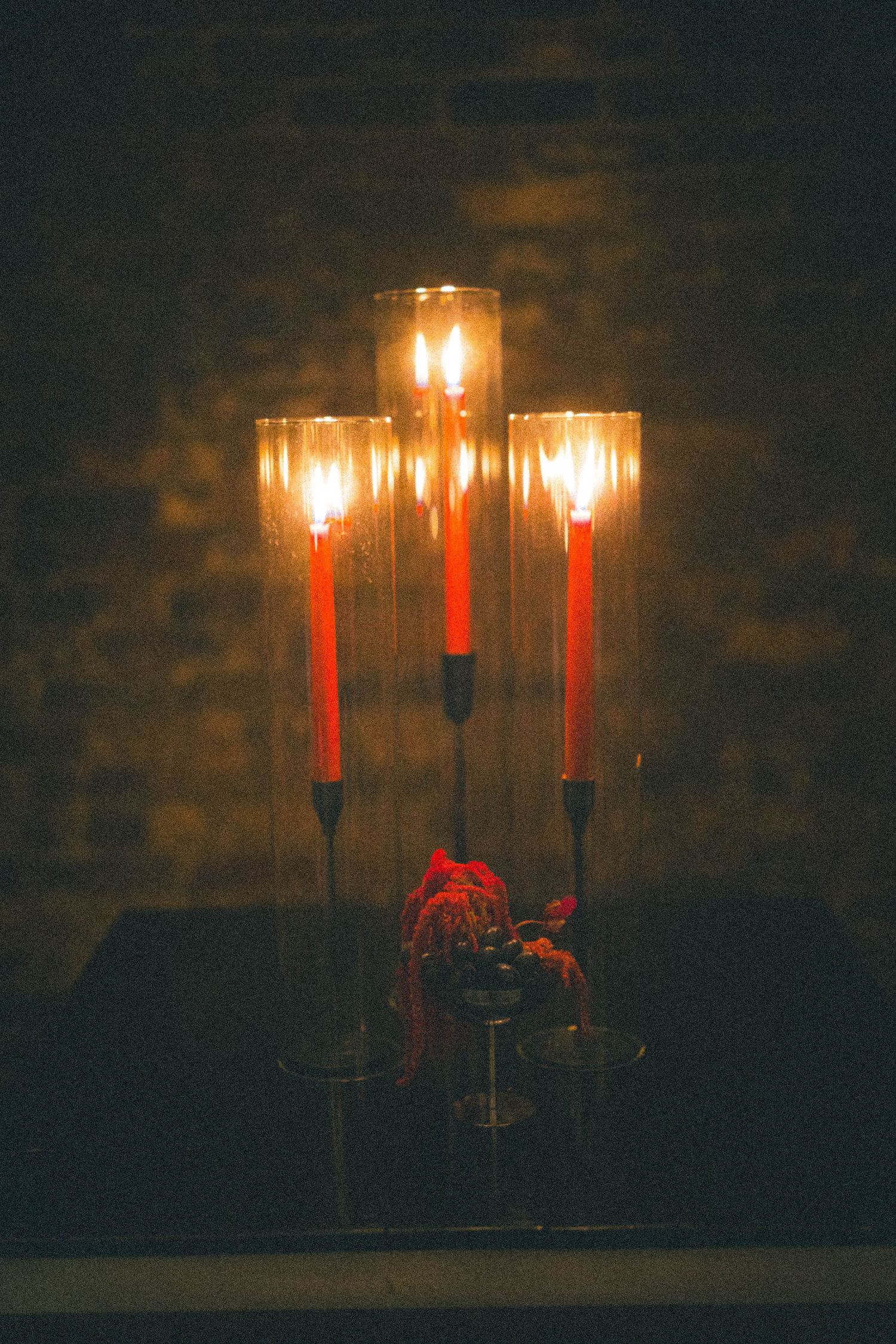 Three red candles in glass hurricane holders glowing warmly on dark table with floral arrangement.