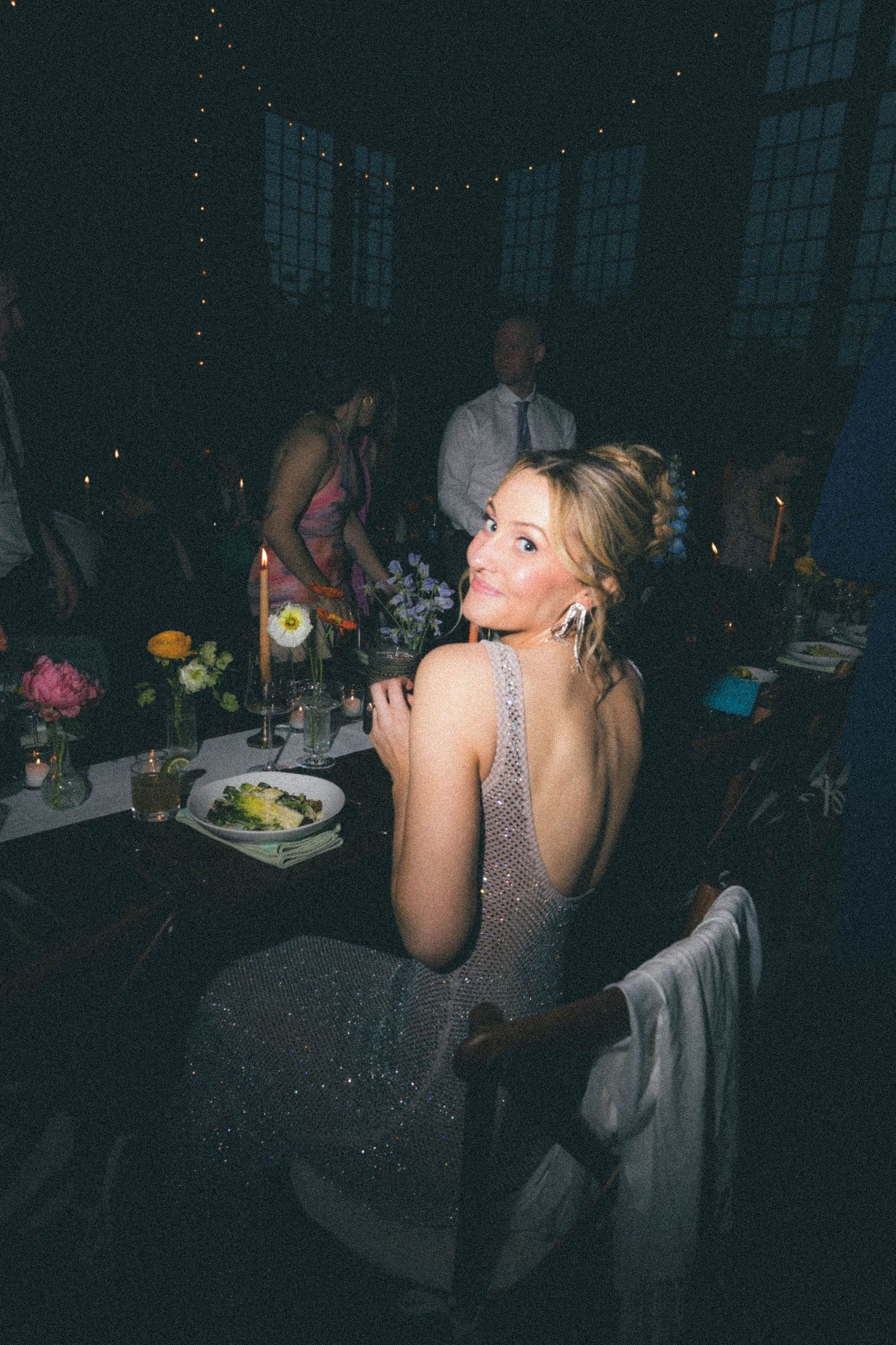 Elegant woman in backless sequin gown smiles at a candlelit wedding reception dinner table.