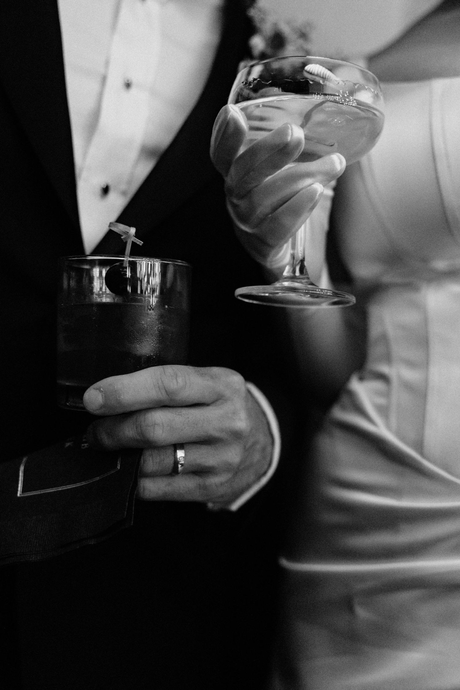 Black and white close-up of bride and groom holding cocktail glasses with wedding rings visible.