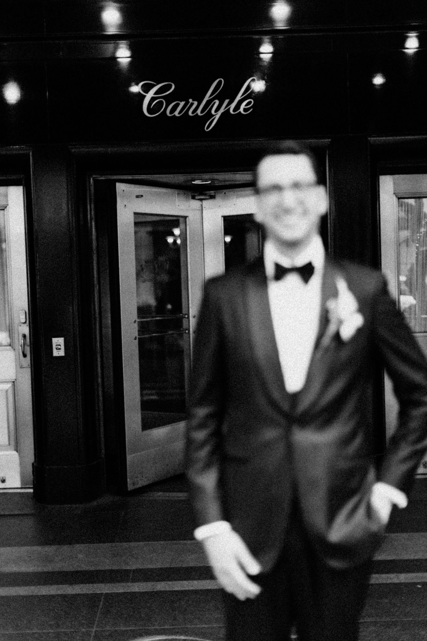 Black and white photo of smiling groom in tuxedo standing outside the Carlyle venue entrance.