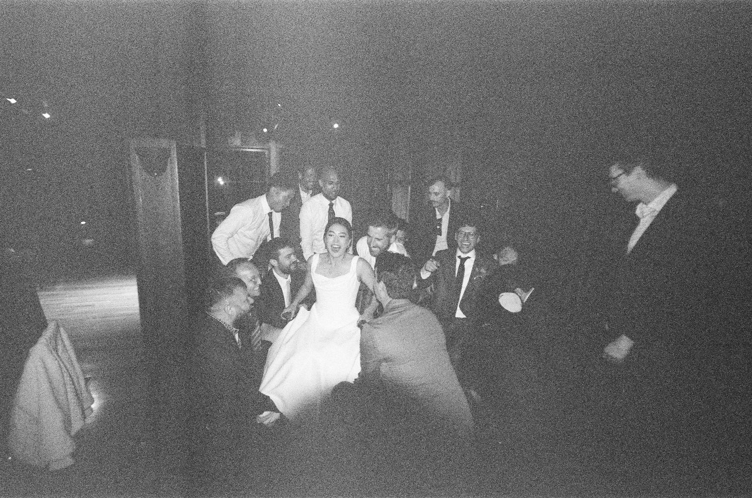 Grainy black and white film photo of bride surrounded by wedding guests gathered together on dance floor.