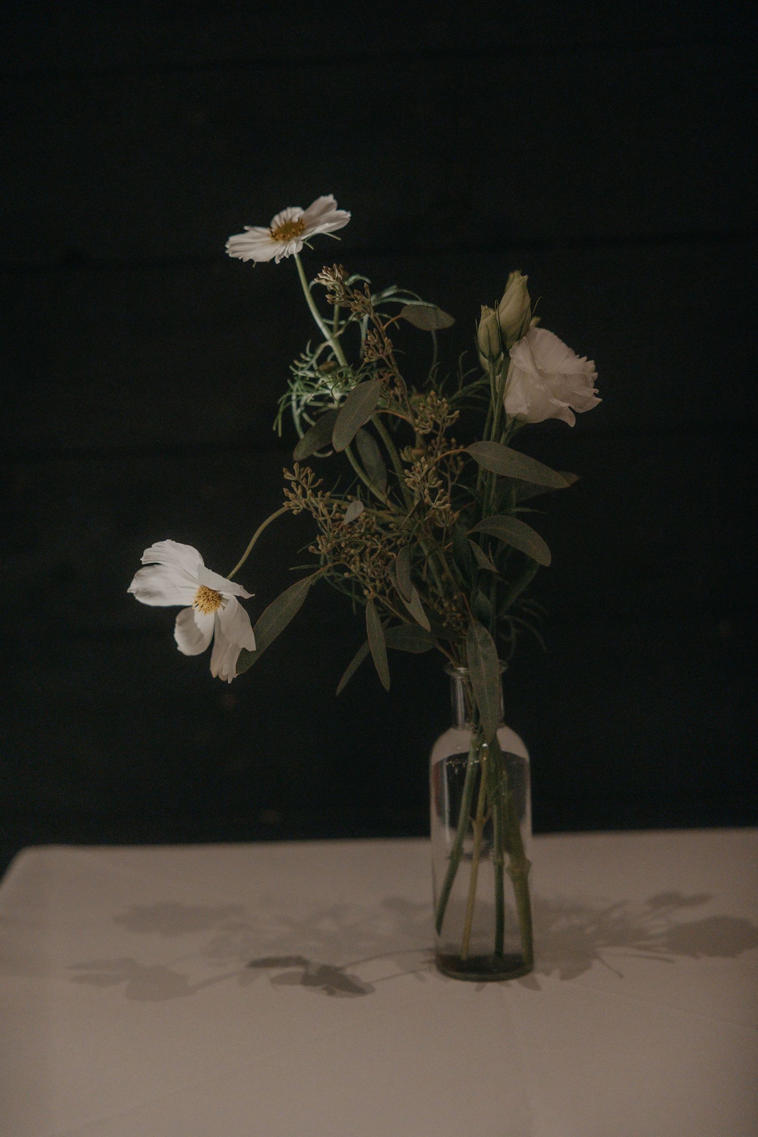 White cosmos flowers and a lisianthus bloom with green foliage in a small glass bottle vase on a white table against a dark background.