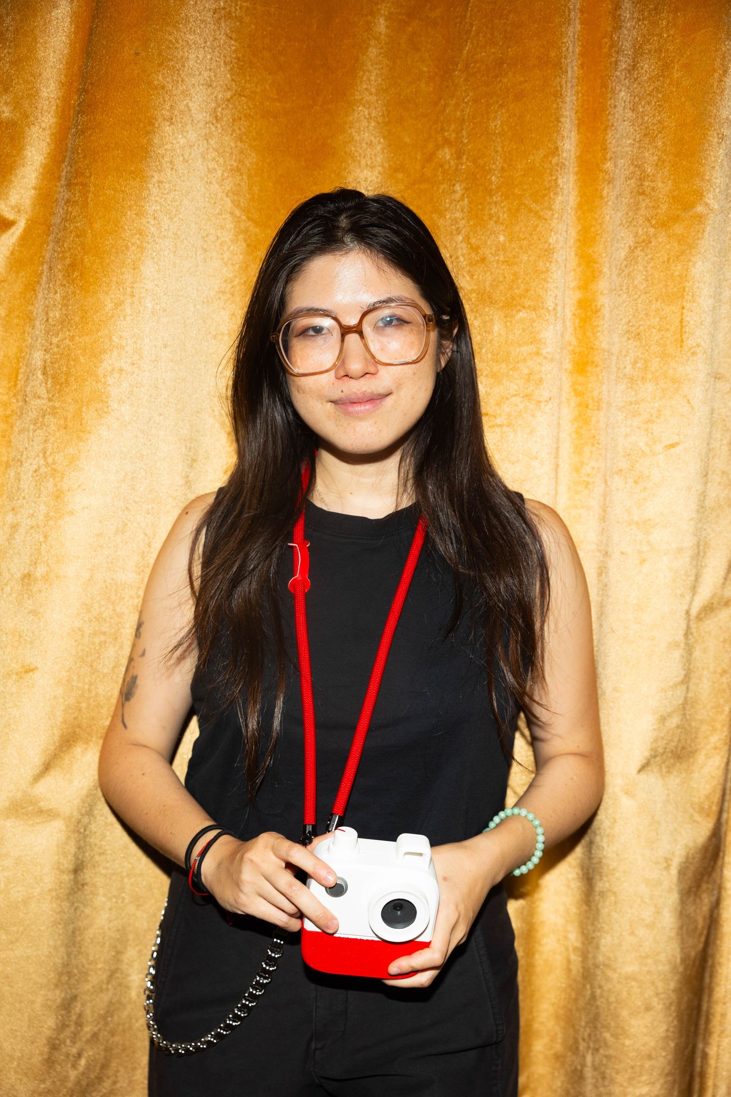 Smiling woman with long dark hair and round glasses holds a white and red instant camera in front of a gold curtain.