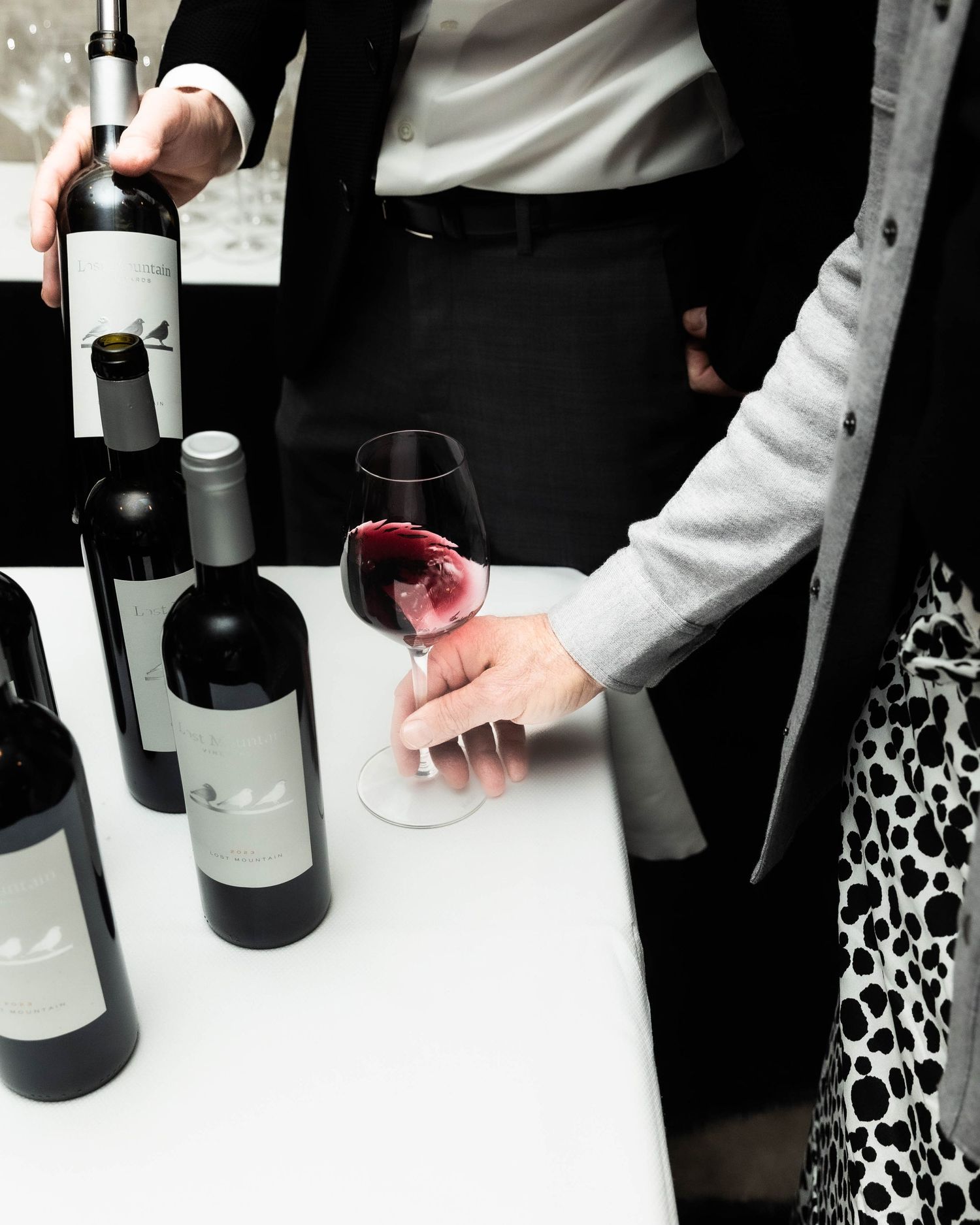 A person swirls red wine in a glass beside several wine bottles on a white tablecloth while another person holds a bottle.