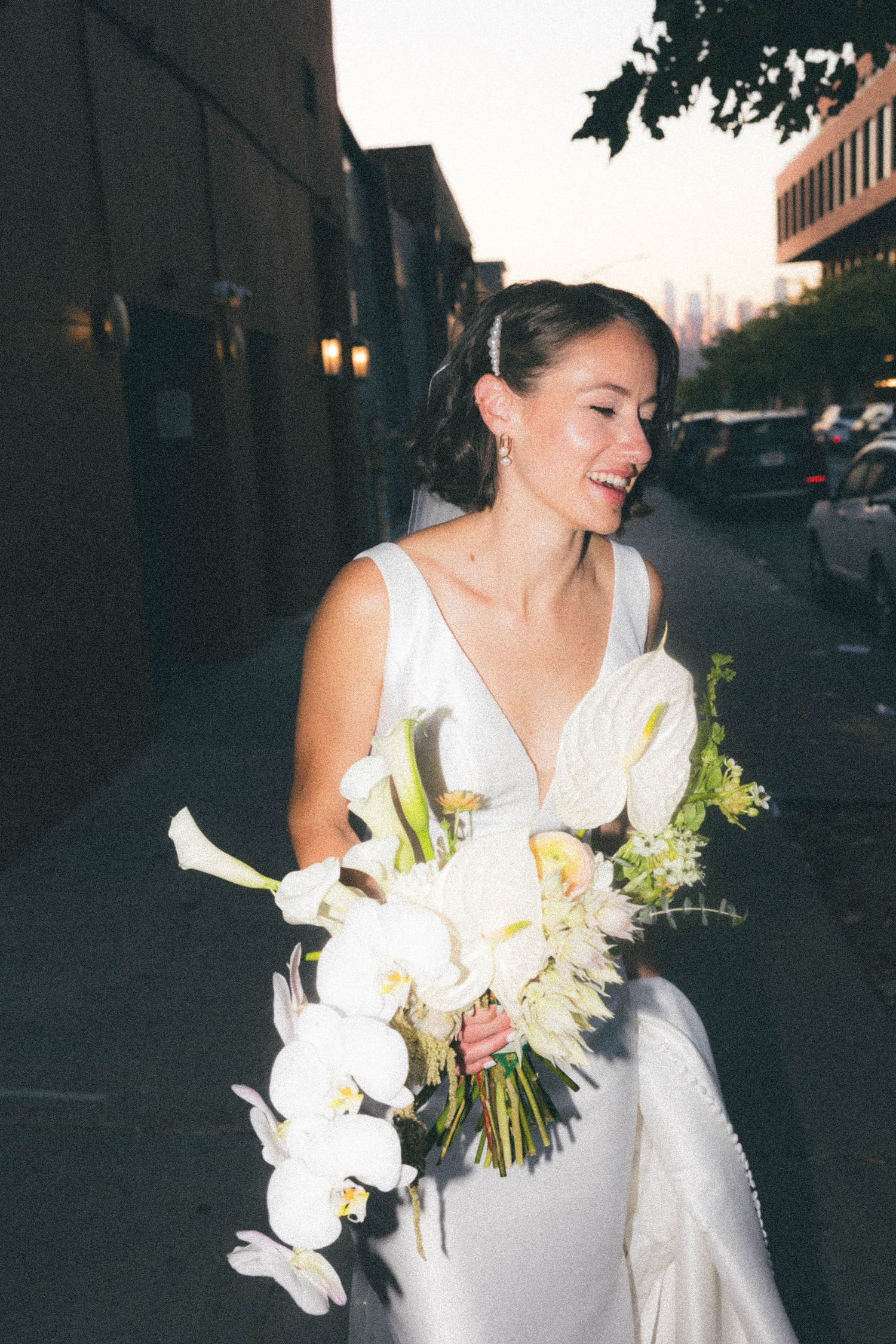 Joyful bride in white gown laughing while holding large white orchid and anthurium bouquet on city street.