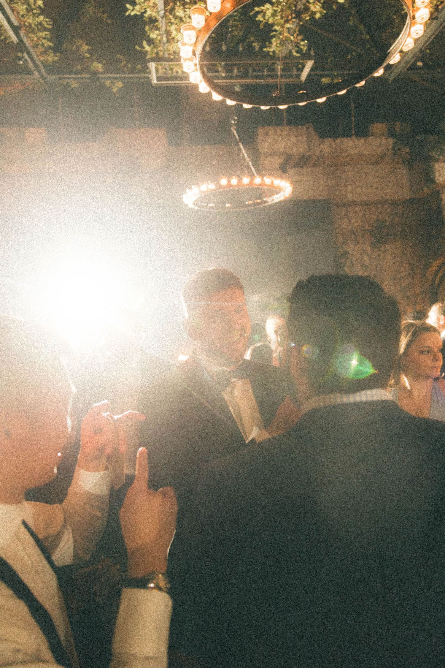 Groom in tuxedo mingling with guests under chandelier at wedding reception with lens flare.