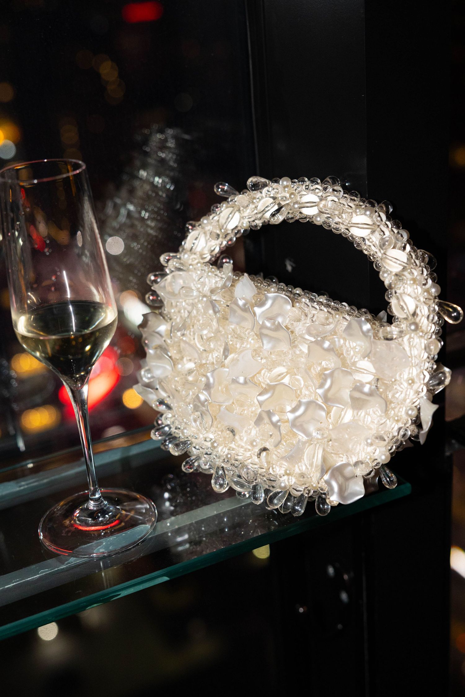 Elegant white beaded bridal purse displayed on glass shelf beside champagne flute at wedding reception.