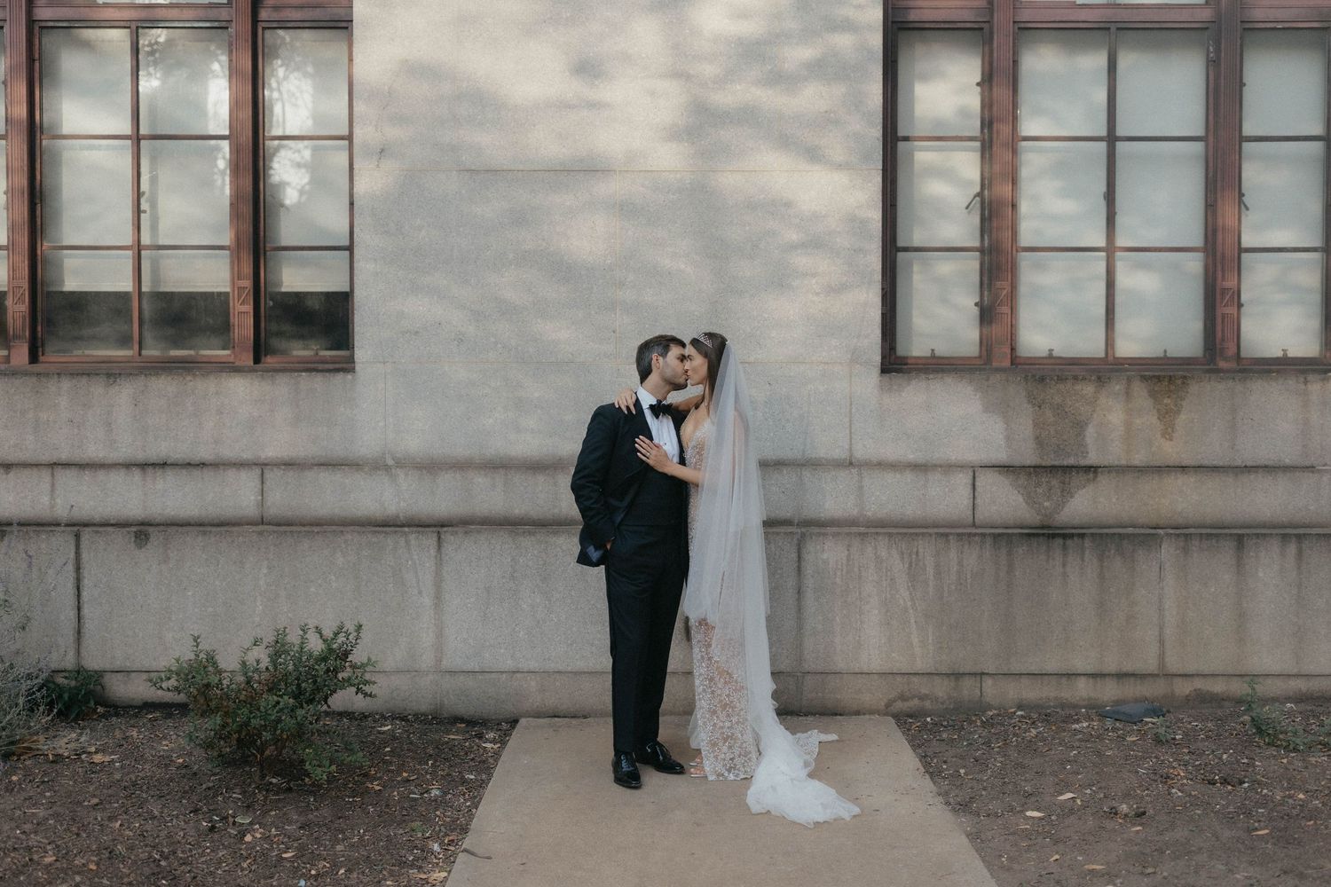 Bride and groom share a tender kiss outdoors against a white concrete wall, her veil flowing gracefully.