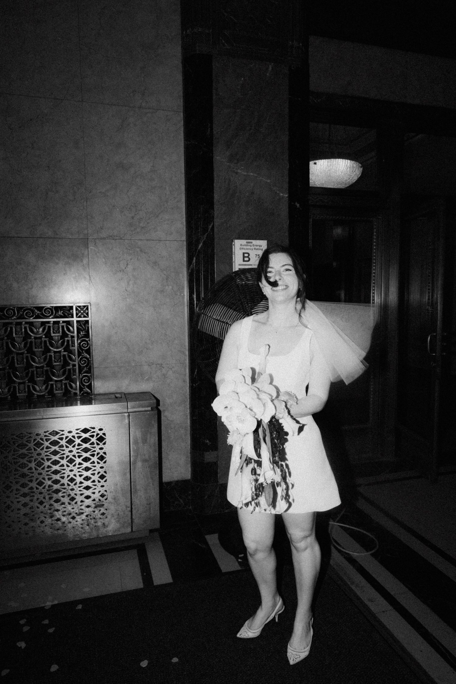 Black and white photo of a smiling bride in a short white dress holding a bouquet indoors.