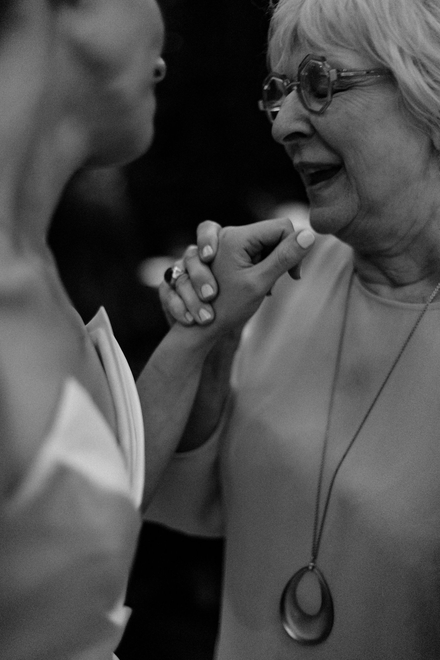 Elderly woman with glasses shares tender moment holding hands with bride at wedding celebration, black and white.