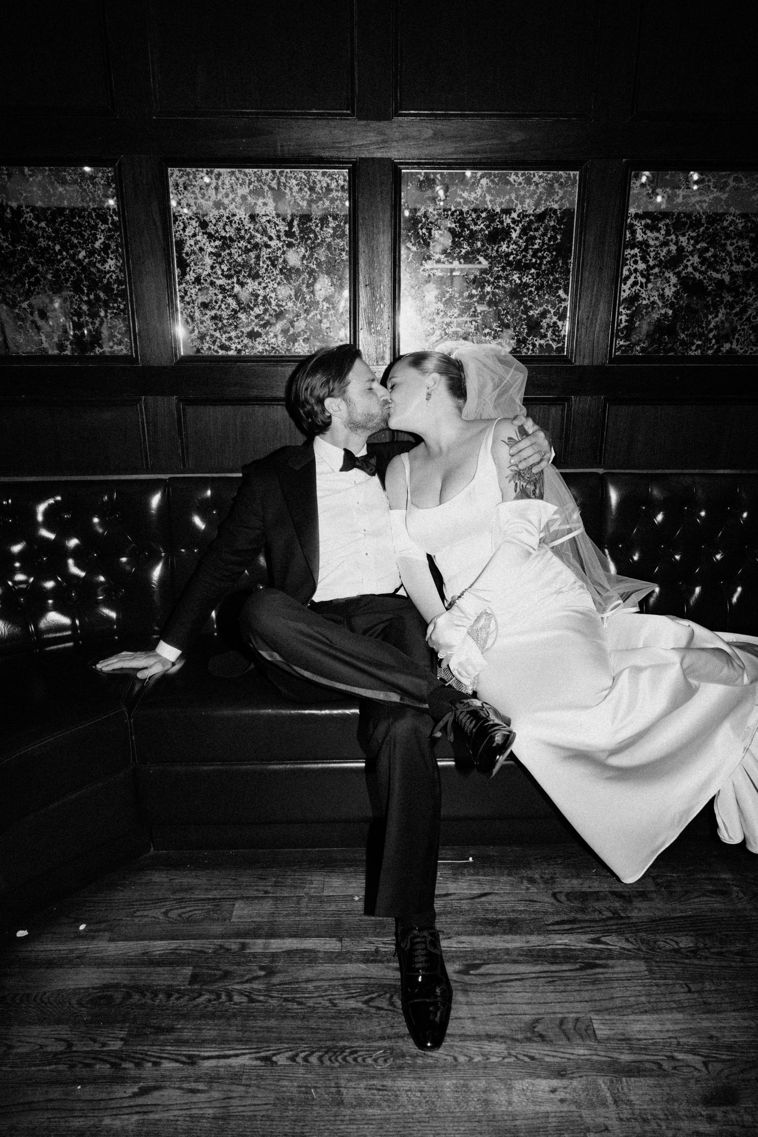 Black and white photo of bride and groom kissing on leather booth at wedding reception.