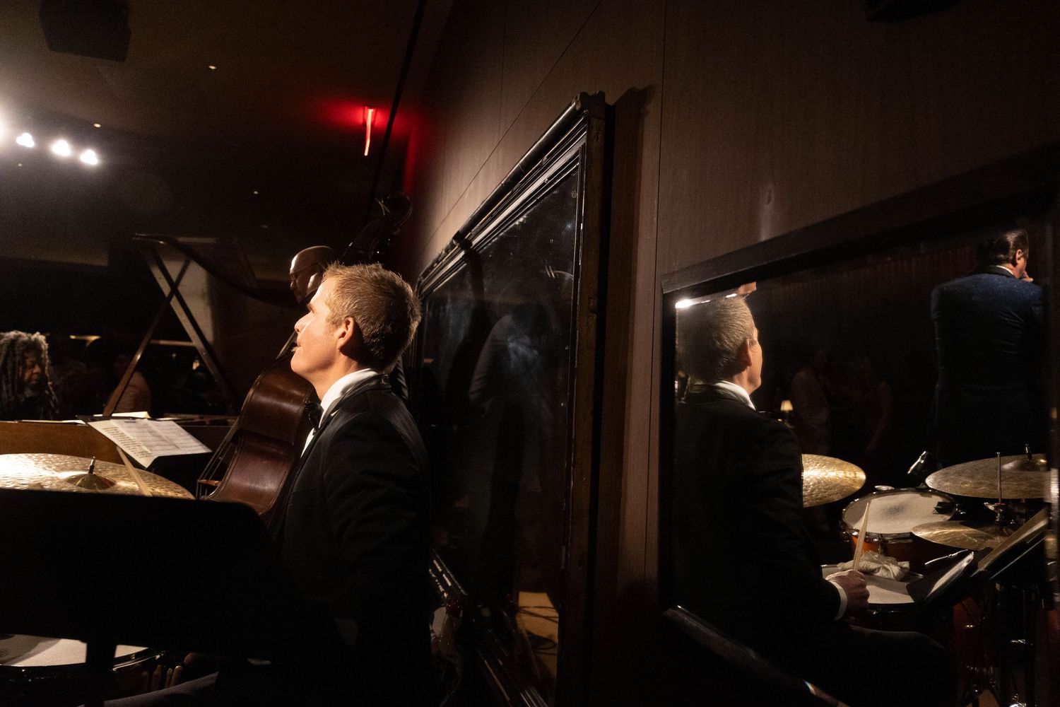 Jazz trio performing in a dimly lit club, with a pianist in a suit, a double bassist, and a drummer reflected in a mirror.