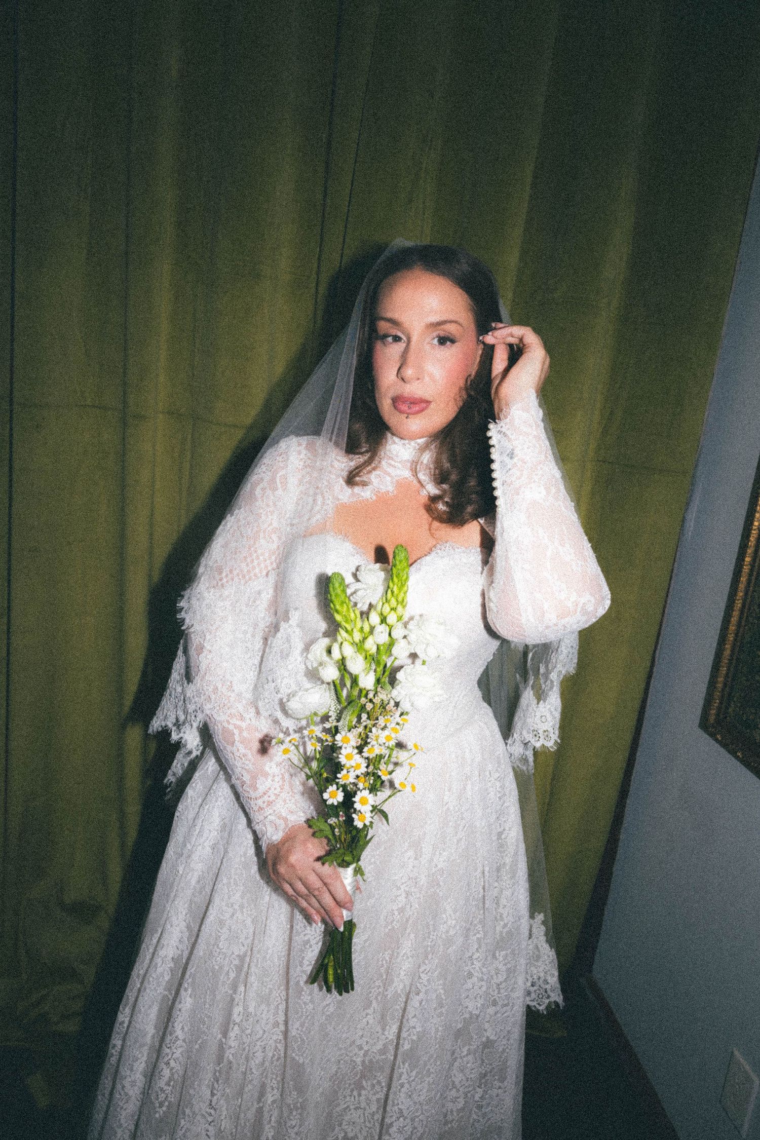 Bride in vintage white lace wedding gown with veil holding wildflower bouquet against green curtain backdrop.