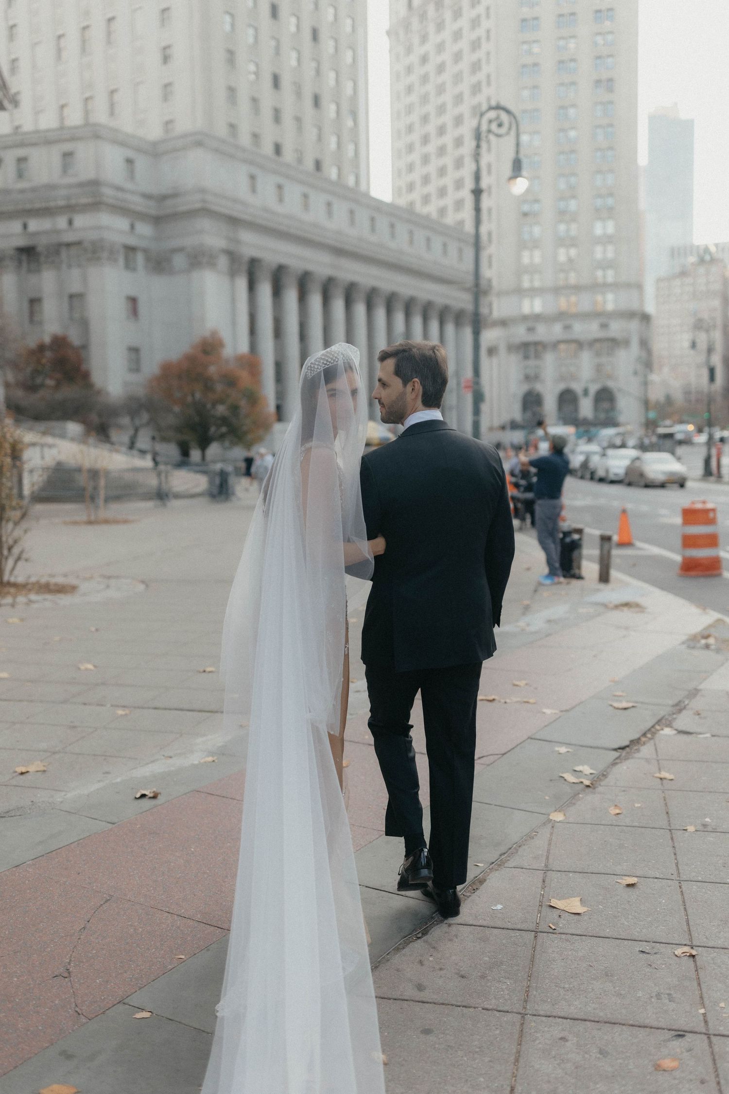 Bride in cathedral veil and groom in black suit walk together past grand neoclassical building in NYC.