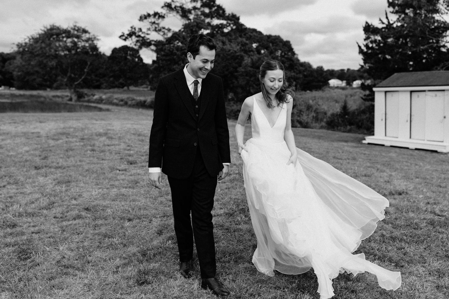 Black and white photo of bride and groom walking hand in hand across green field at outdoor wedding.