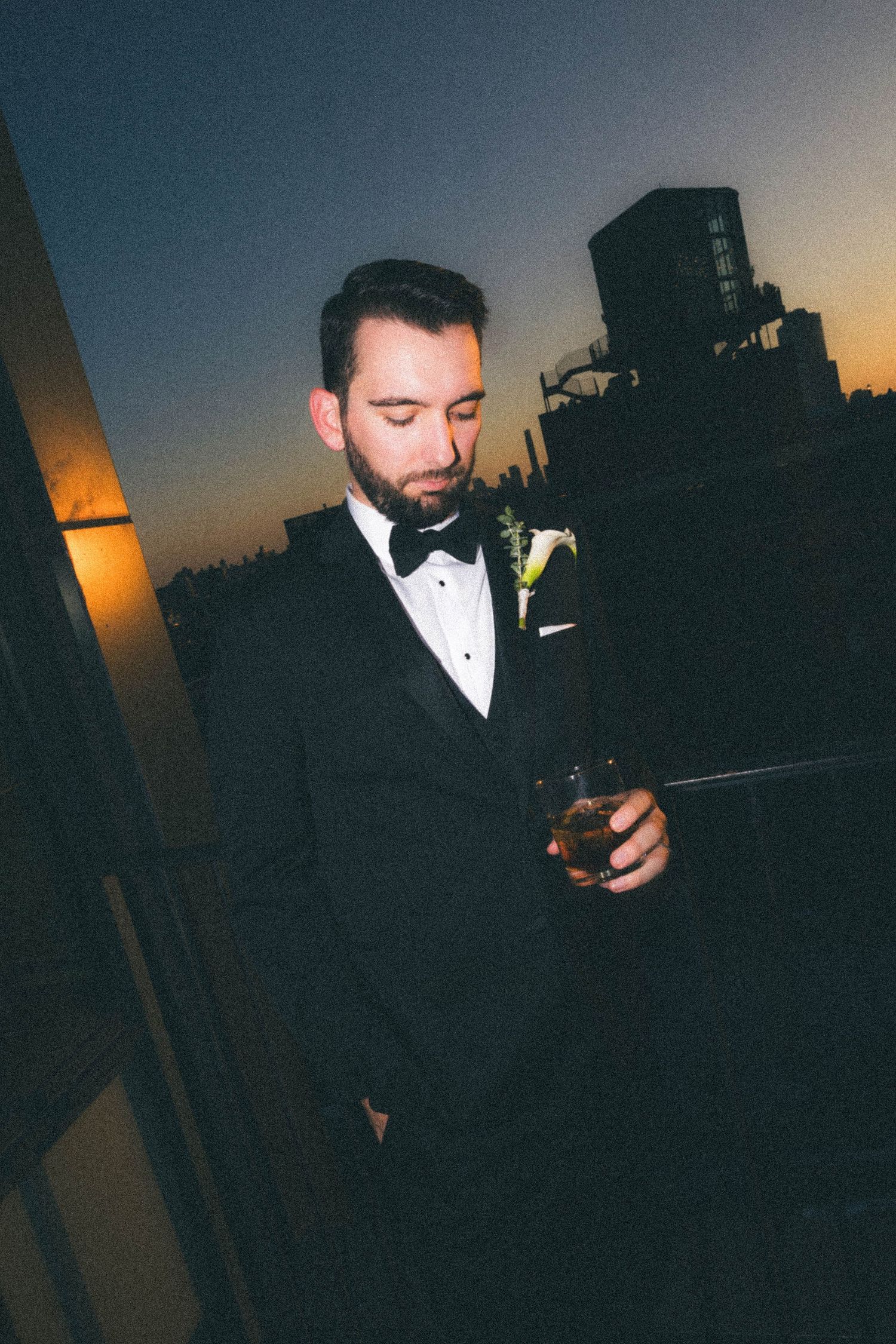 Groom in black tuxedo holding whiskey glass on rooftop at sunset with city skyline behind him.
