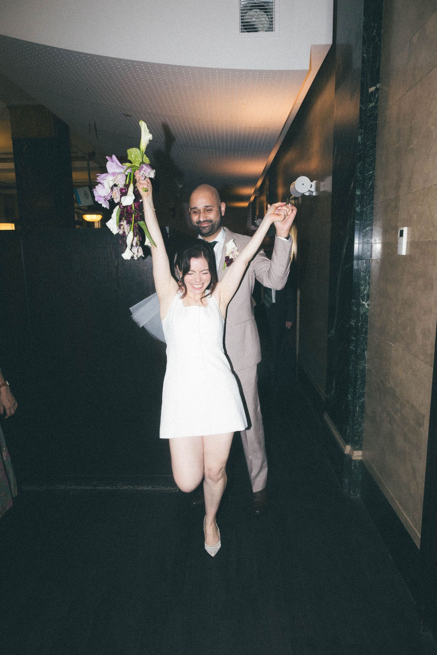 Excited bride and groom walking down hallway with arms raised holding floral bouquet in celebration.