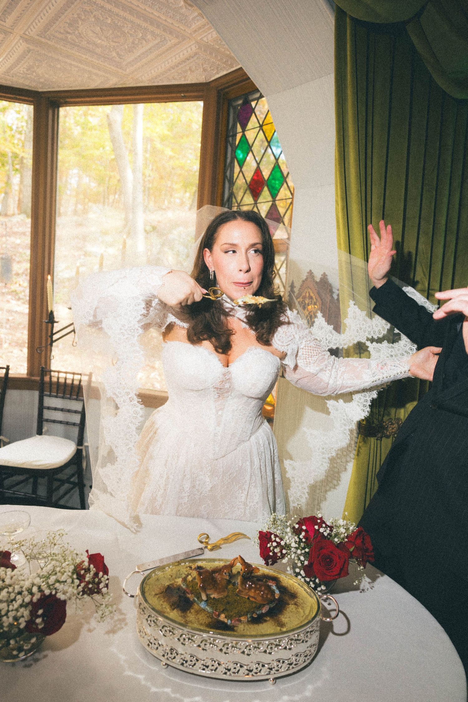 Bride in white lace gown with veil reacting dramatically near wedding cake table with red roses.