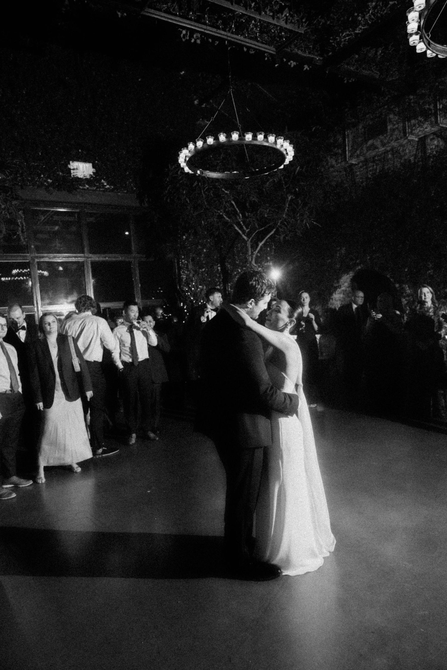 Black and white first dance photo of bride and groom under chandelier at wedding reception.