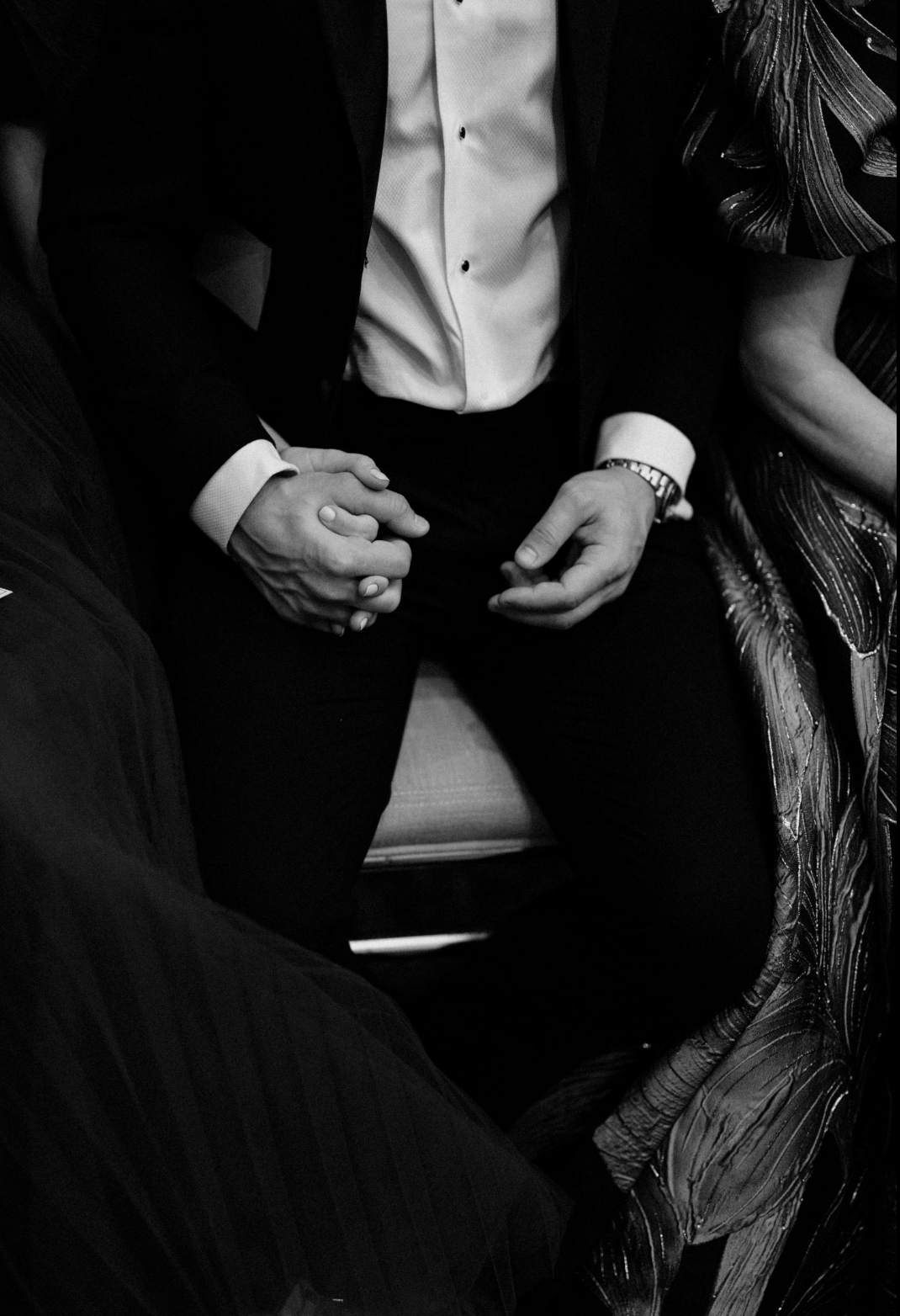 Black and white close-up of groom in tuxedo with clasped hands and bracelet seated during wedding ceremony.