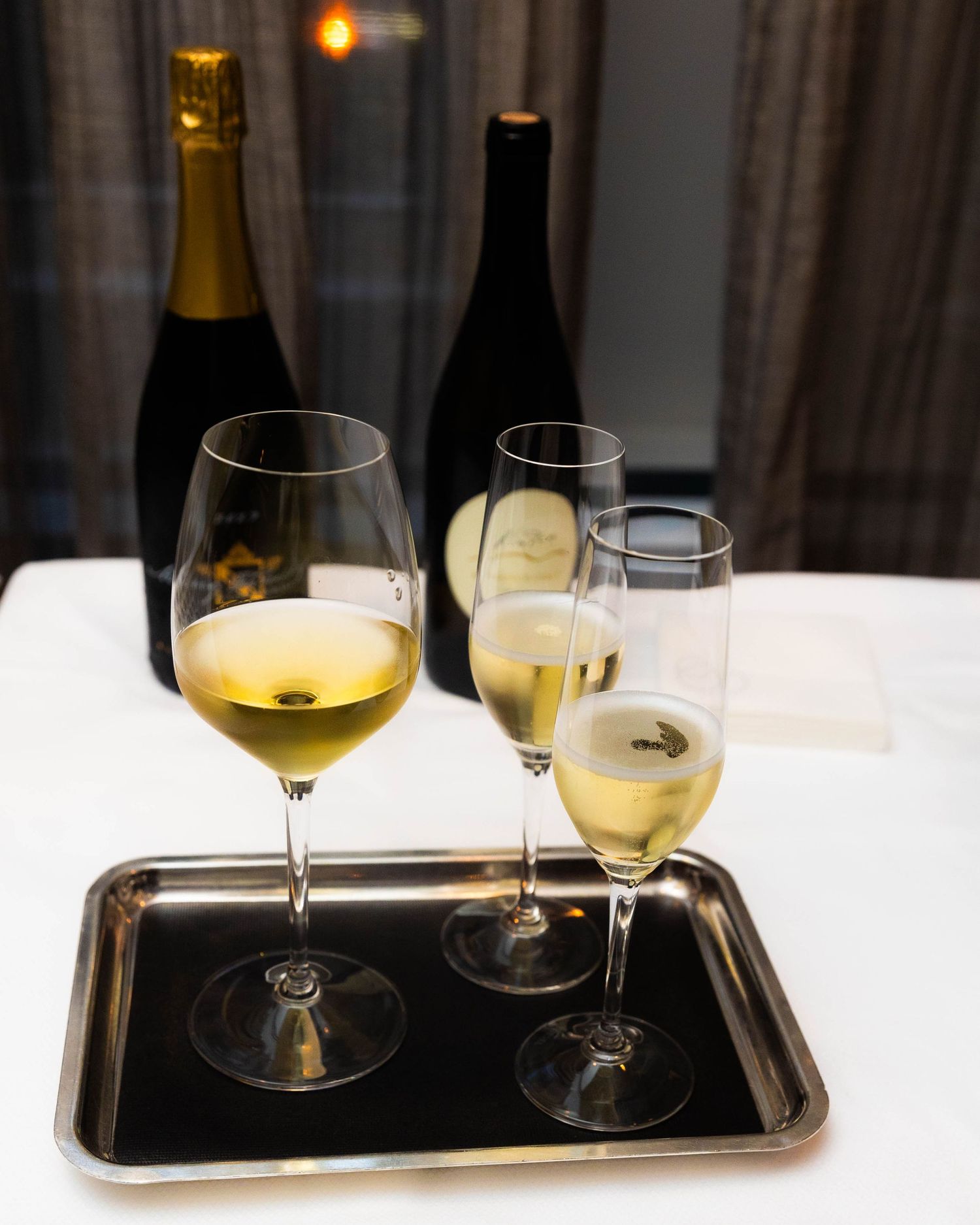 Three glasses of sparkling white wine on a black serving tray, with two champagne bottles standing on a white-clothed table behind.