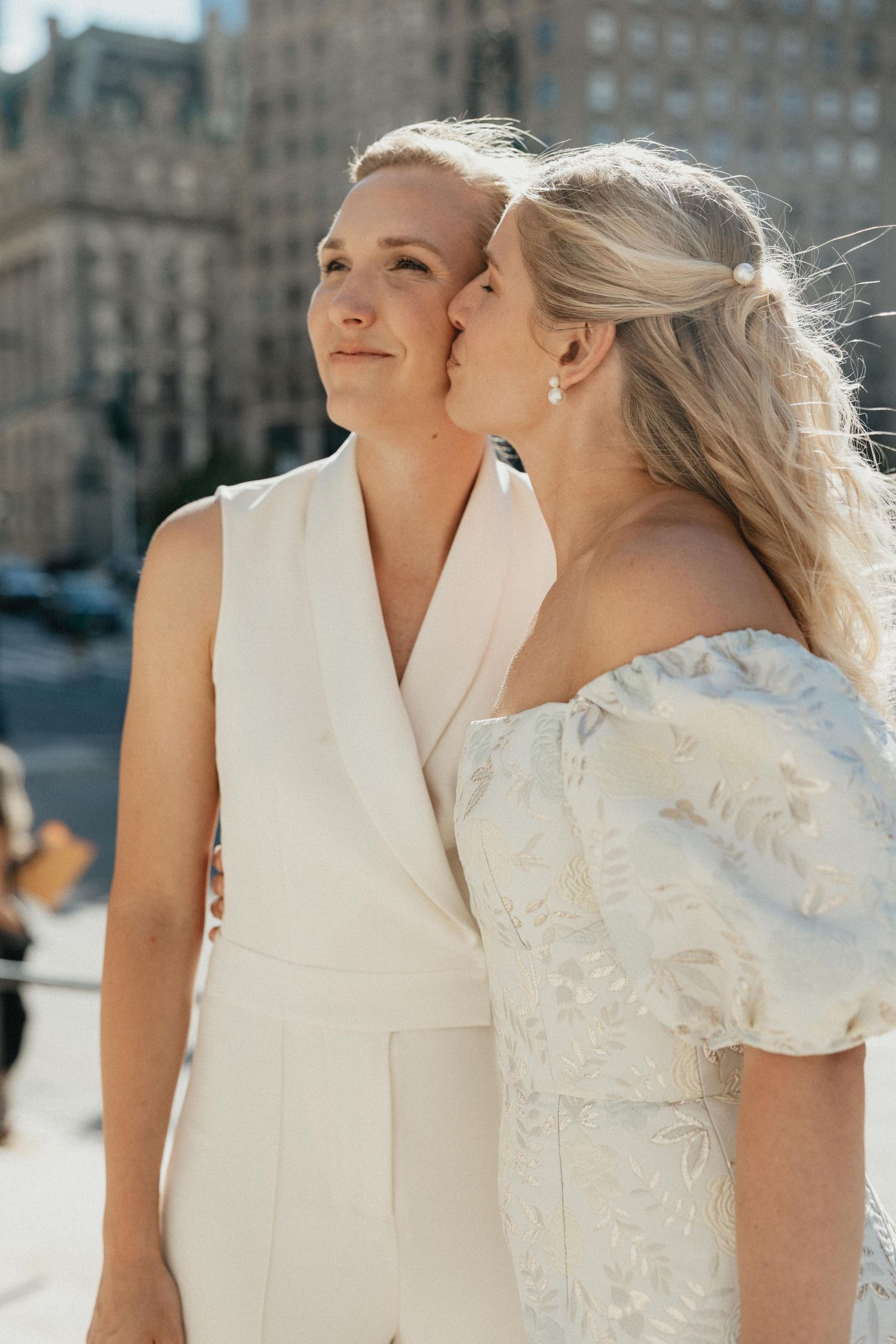 Two brides outdoors in sunlight, one kissing the other on the cheek in a city setting.
